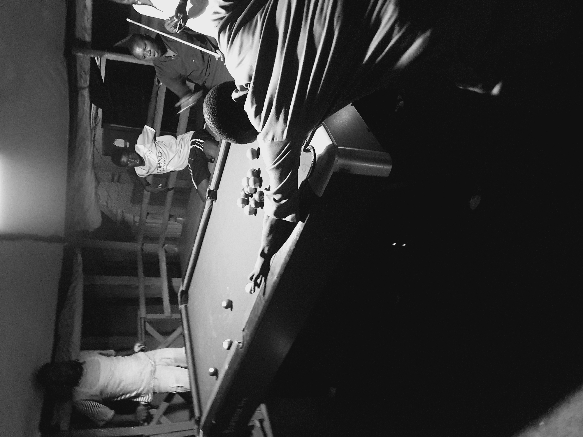 The black and white image depicts a group of young men playing pool inside a dimly lit, rustic structure that appears to be outdoors or semi-enclosed. The environment suggests a casual, possibly makeshift, recreational area. The pool table is the central focus, with several billiard balls arranged on its surface.
In the foreground, a young man is bent over the pool table, cue in hand, preparing to strike a ball. His body is angled towards the table, and his focus is clearly on the game. His clothing is dark and appears to be a loose-fitting shirt.
Further back, several other individuals are visible. One young man in a light-colored t-shirt with "ETIHAD AIRWAYS" visible is sitting or leaning near the table. Another man, wearing a dark shirt, is holding a pool cue and looking towards the table. A third individual, partially obscured, is standing behind the table. In the far background, another person is visible standing with their back to the camera.
The structure's construction is visible, with wooden beams and railings suggesting an open-air or partially covered setting. The lighting is low, creating strong shadows and highlighting certain areas of the scene.
The context of Lokoja, Nigeria, suggests this scene is likely taking place in a neighborhood or community gathering spot. The overall atmosphere is one of leisure and friendly competition.