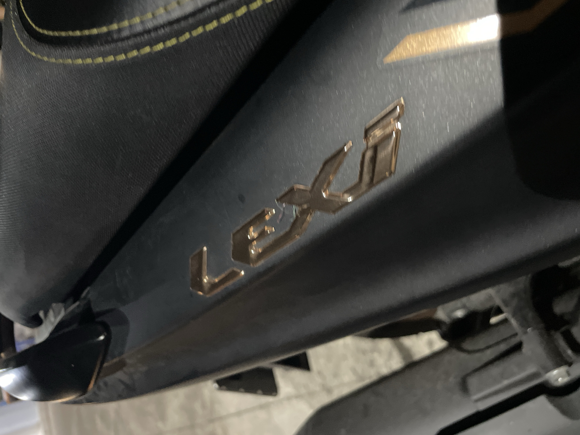 The image shows a close-up, angled view of a dark gray motorcycle's side panel with a gold-colored "LEX" logo. The "L" is partially visible on the left, followed by the "E" and "X" prominently displayed. A black textured seat with yellow stitching is visible in the upper right corner of the frame. The background is blurred and appears to be an indoor setting, possibly a garage or workshop, with concrete flooring and some indistinct objects. There are no people or animals present in the image. The lighting suggests an artificial light source. The scene does not provide any specific indicators that it is located in Bac Nha Trang Ward, Vietnam.