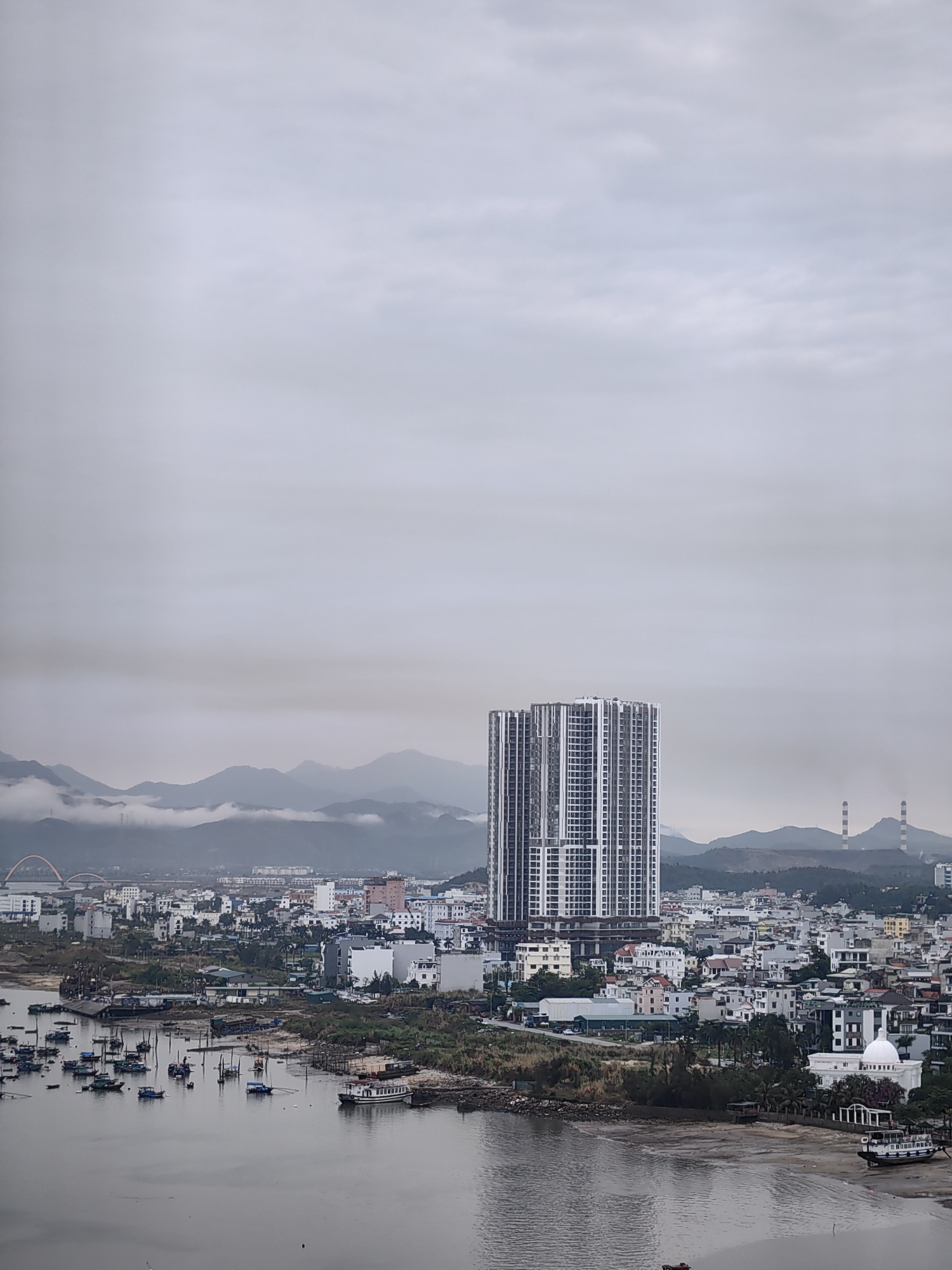 The image depicts a cityscape in Hong Gai Ward, Vietnam, under a cloudy sky. In the foreground, a body of water is visible with numerous small boats anchored near the shore. The middle ground features a dense collection of buildings, ranging in height from low-rise houses to a prominent high-rise apartment complex in the center of the frame. A mosque-like building with a white dome can be seen on the right side of the image. In the background, mountains are partially obscured by mist. Two tall smokestacks are visible on a distant hill. A curved bridge is visible near the left edge of the frame. There are no people visible in the image.