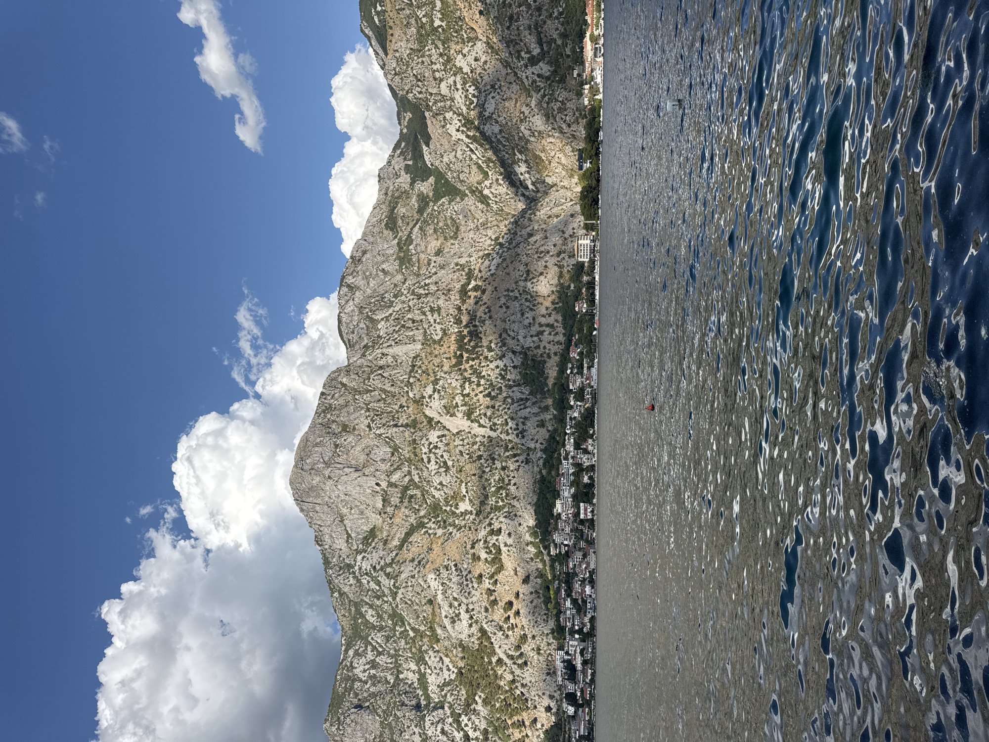 The image depicts a scenic view of Muo, Montenegro, from the water. In the foreground, the dark, rippling water of a bay reflects the sky and sunlight, creating patterns of light and shadow. A small red buoy floats in the middle distance on the water. Behind the water, a coastal town with white- and light-colored buildings is nestled at the foot of a massive, rugged mountain range. The mountains are characterized by their rocky, arid slopes, with patches of green vegetation in the lower sections. The sky above is a vibrant blue, dotted with fluffy white cumulus clouds. The overall impression is one of a serene, picturesque coastal landscape with a prominent mountainous backdrop.