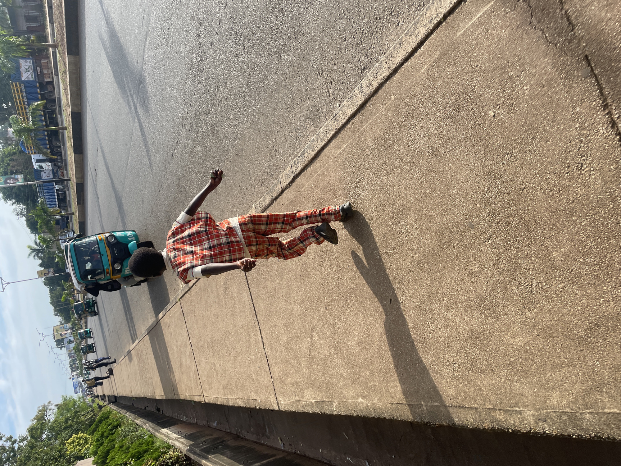 A young boy is walking on a concrete sidewalk next to a busy road in Jos, Nigeria. The boy is wearing a red and white plaid shirt and matching plaid pants, and he is looking away from the camera. On the road, there are several vehicles, including a turquoise tuk-tuk and a large blue truck. The road is lined with palm trees and other greenery. The sky is cloudy, and the sun is casting long shadows on the sidewalk. The overall scene depicts a typical urban environment in Nigeria.