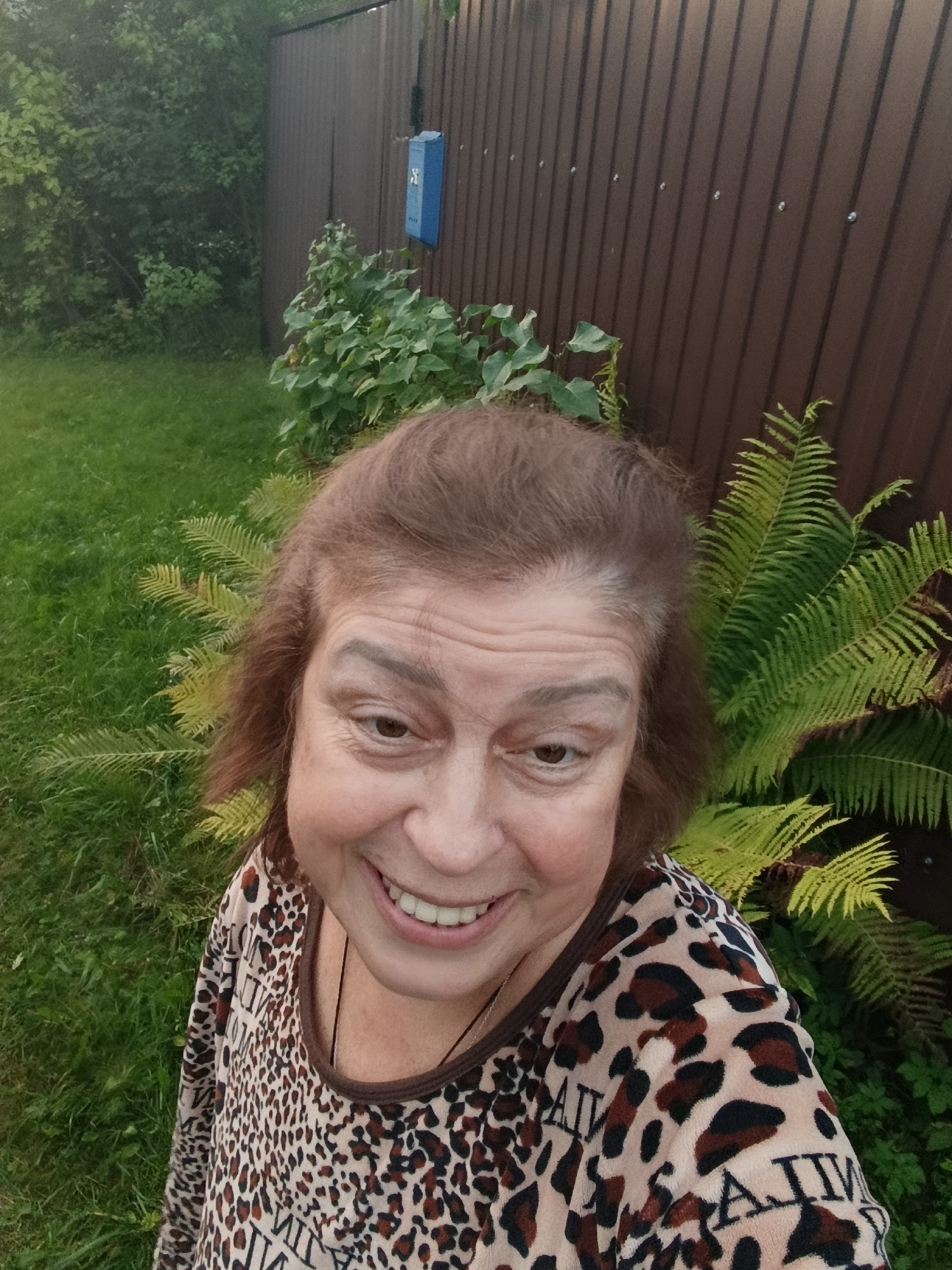 A selfie shows a middle-aged woman with brown hair and smiling, looking at the camera. She is wearing a long-sleeved, leopard-print shirt. Behind her, there is a brown corrugated metal fence, green grass, and some green plants. A blue mailbox is attached to the fence. The scene appears to be an outdoor backyard in Veriya, Russia.