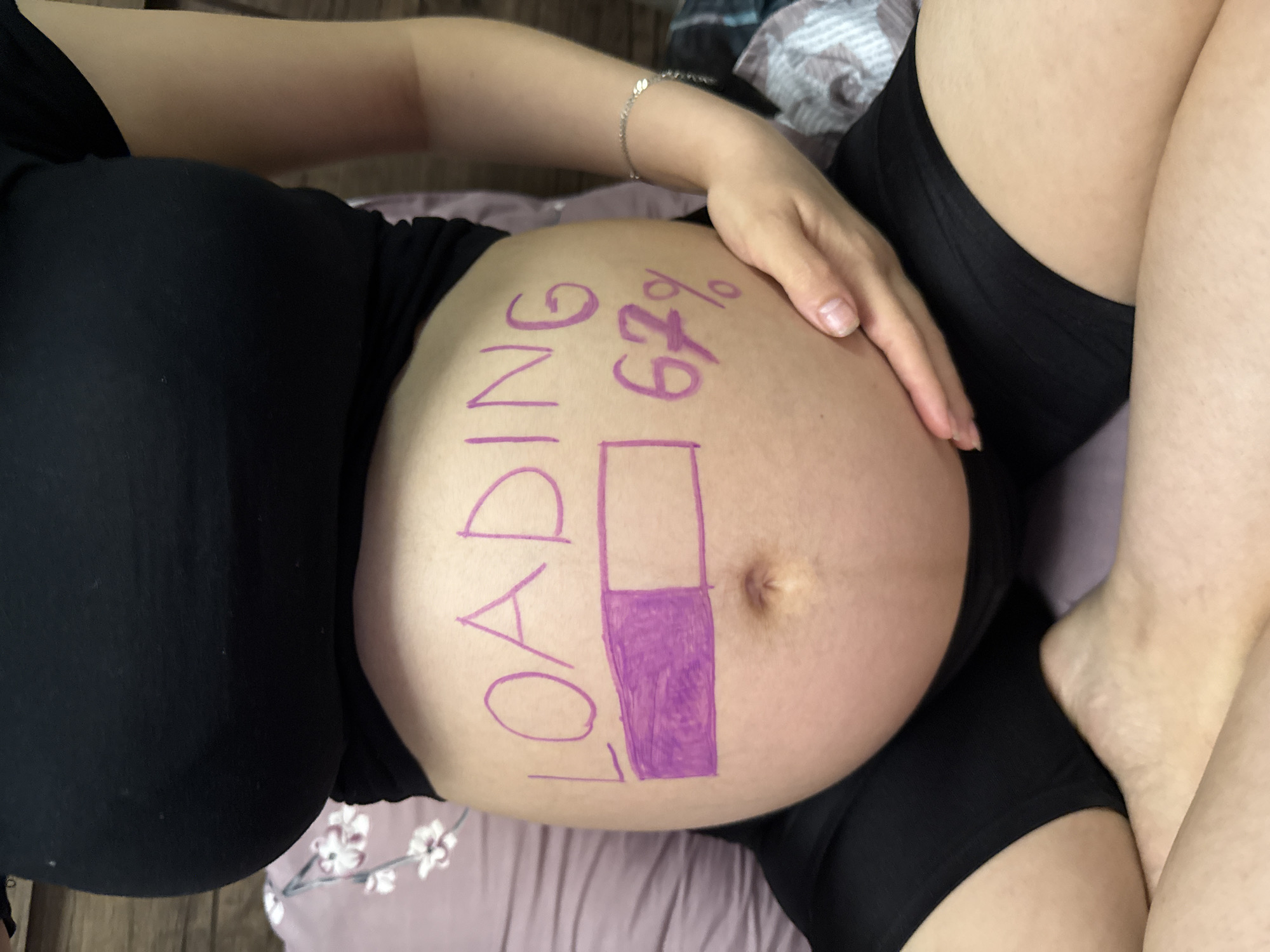 The image shows a close-up of a pregnant person's belly with "LOADING" and a progress bar drawn on it in purple marker. The progress bar is partially filled, indicating 67% completion. The person is wearing a black top and black shorts. They are sitting, with their hands resting on their belly. The background consists of a wooden wall and pillows. The image is taken indoors. There is no information in the image to suggest a location in Tbilisi, Georgia.
