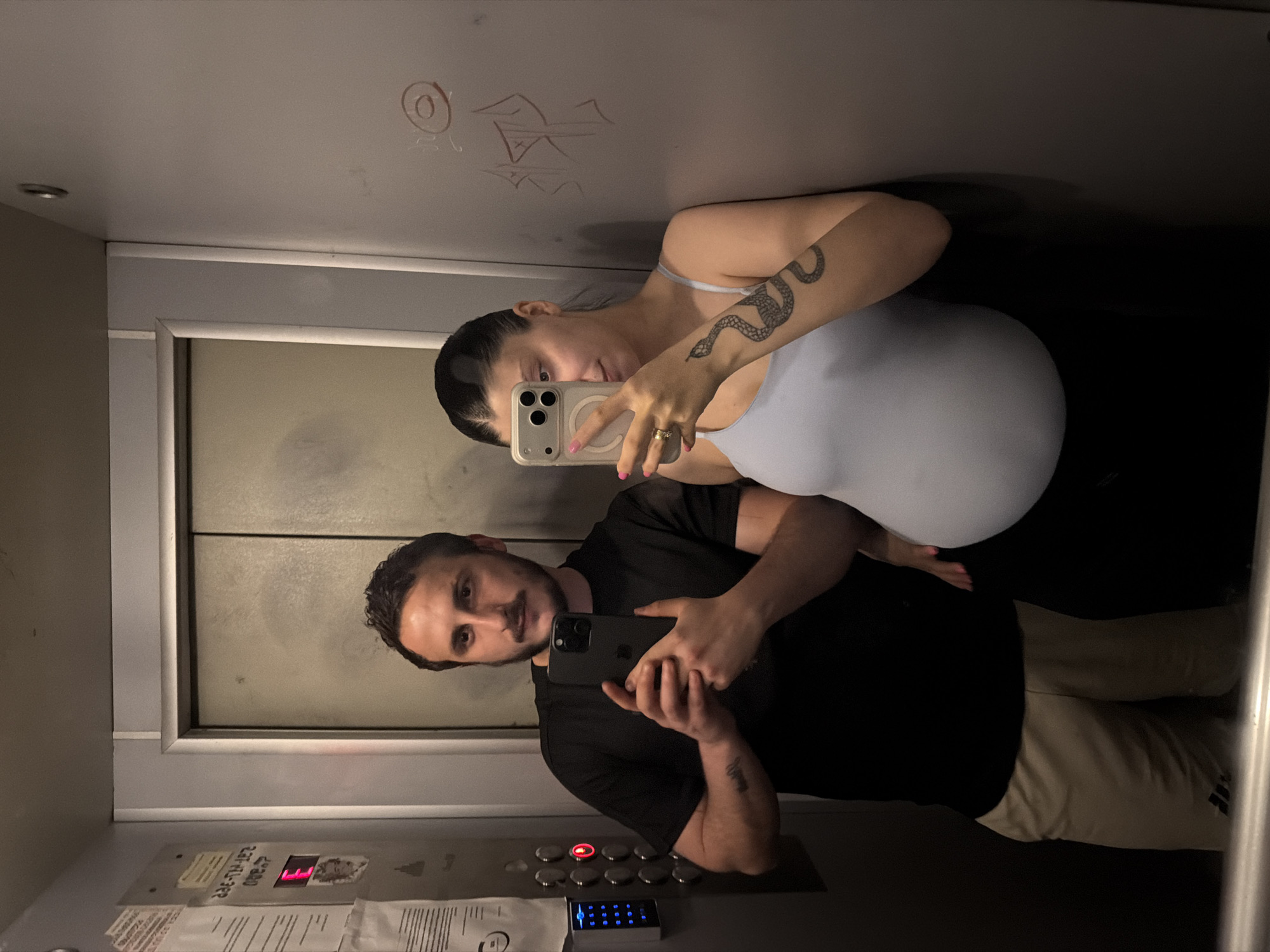In an elevator in Tbilisi, Georgia, a couple poses for a mirror selfie. The man, with dark hair and a mustache, wears a black t-shirt and tan shorts, holding a smartphone to take the photo. The woman, with dark hair pulled back, wears a light blue tank top and black pants, also holding a smartphone. Her pregnant belly is prominent. She has a tattoo of a snake on her left arm. The elevator walls are gray, with some graffiti visible. An elevator control panel with buttons and a digital display showing the letter "E" is to the left of the couple.