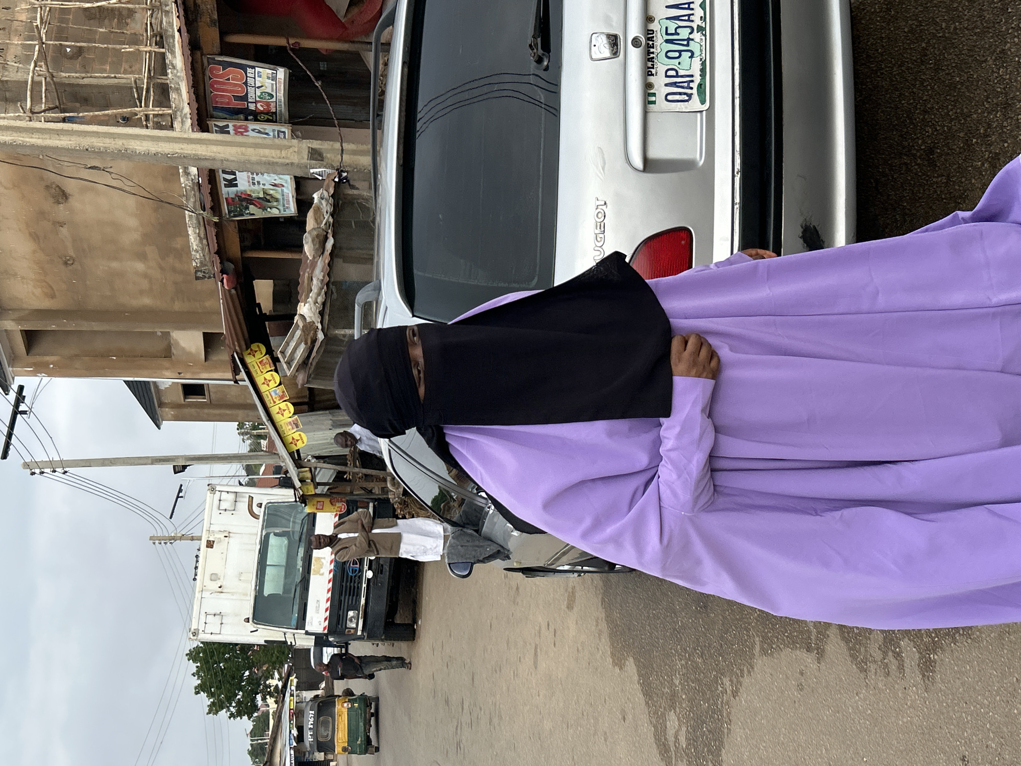 A woman wearing a long purple gown and a black veil stands next to a silver Peugeot car with a Nigerian license plate "QAP-945AA" registered to Plateau state. The location appears to be a street in Jos, Nigeria, with buildings and utility poles visible in the background. A white truck and other vehicles are further down the street, including a yellow and green tricycle. Several people are visible in the background. The foreground shows a section of a paved street.
