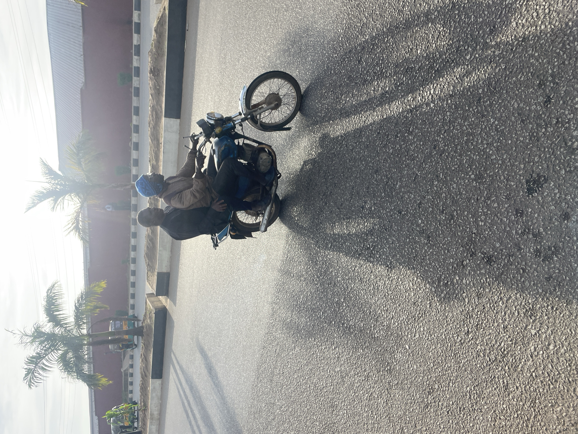 The image depicts a street scene in Jos, Nigeria. Two individuals are riding on a blue motorcycle, with the driver wearing a blue headscarf and a light brown jacket, and the passenger wearing a dark top. They are moving from left to right across the frame. The motorcycle has a visible exhaust pipe, front wheel, and handlebars. In the background, palm trees line the street, and a red-walled building with a corrugated metal roof is visible. There are also some utility vehicles parked on the side of the road. The ground is asphalt, with tire tracks and shadows of the motorcycle and trees cast upon it. The lighting suggests it is either early morning or late afternoon.