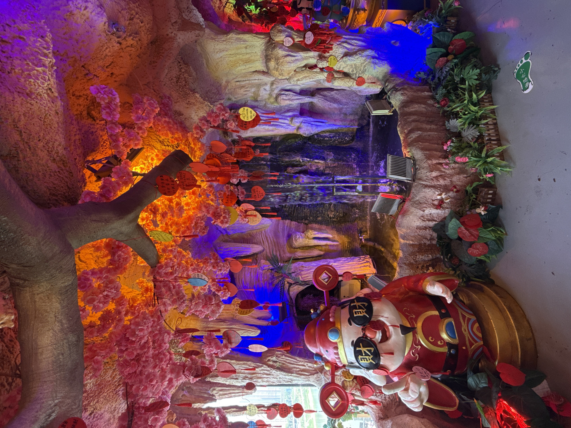This image captures an elaborate indoor decorative display, likely within a themed restaurant, shopping mall, or cultural attraction in Yuzhong District, China. The scene is rich with artificial elements designed to evoke a festive and natural atmosphere.
In the foreground, a large, colorful statue of a chubby, red-robed figure, reminiscent of a Chinese God of Wealth (Caishen), is prominently featured. The figure wears dark glasses with gold Chinese characters: "招财" (attract wealth) on one lens and "进宝" (bring in treasures) on the other, forming the common idiom "招财进宝" (ushering in wealth and prosperity). It holds a traditional Chinese coin.
To the left of the statue is an artificial tree with a thick trunk, heavily adorned with pink, cherry blossom-like flowers. Numerous small, heart-shaped or circular red tags, some with indiscernible Chinese characters, hang from the branches. Further into the scene, artificial rock formations are illuminated by vibrant blue and orange lights, creating a grotto-like effect that suggests a waterfall or pond area. More decorative tags and artificial green foliage are scattered across these rock structures.
The overall activity is the viewing or enjoyment of this static, decorative installation. The bright, colored lighting contributes to a festive and somewhat fantastical mood. On a grey wall in the upper right corner, a green footprint sticker with the Chinese characters "小心地" (carefully / mind your step) is visible. Some yellow circular tags also appear, one showing a partial phone number "835-816" and other small characters. The display is intricately designed, blending cultural symbolism with artificial natural beauty.