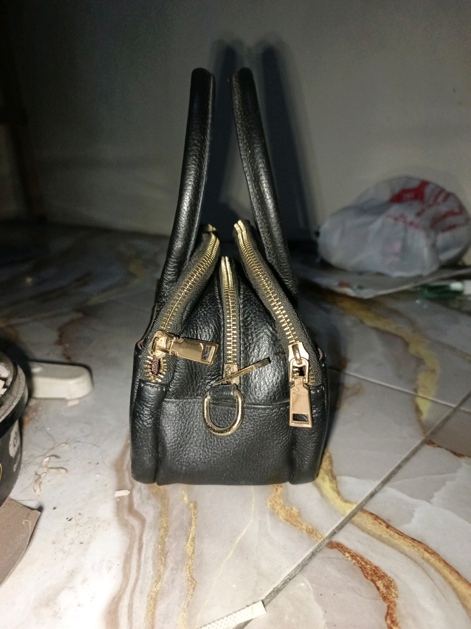 A black leather handbag with gold zippers is positioned upright on a tiled floor. The handbag has two top handles and appears to be empty. In the background, a crumpled plastic bag with red markings is visible. The floor tiles are a marbled pattern of white, brown, and yellow. To the left of the handbag, a portion of a circular object with a metallic edge and dark coloring is visible, along with some debris. The lighting in the scene is dim, creating shadows. The location is identified as Ketegan, Indonesia, though no specific landmarks confirm this.