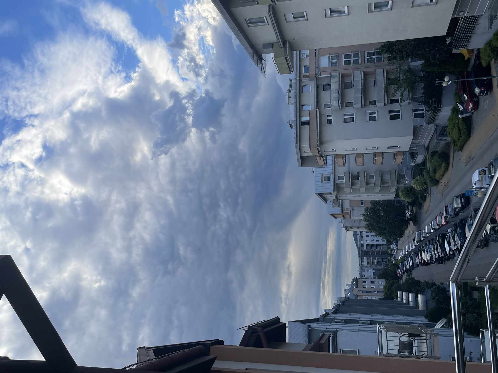 An outdoor, eye-level, vertical, full shot shows a cloudy, gray sky over a residential street lined with apartment buildings and parked cars. The sky is mostly filled with large, dark, textured clouds, with patches of lighter sky and blue sky visible on the right. A dark, angled structure, possibly a balcony railing or awning, is in the upper left corner. The apartment buildings are modern, with light-colored facades, balconies, and numerous windows. On the street, a long line of cars is parked on both sides. The overall scene suggests a residential neighborhood under a somewhat overcast sky.
