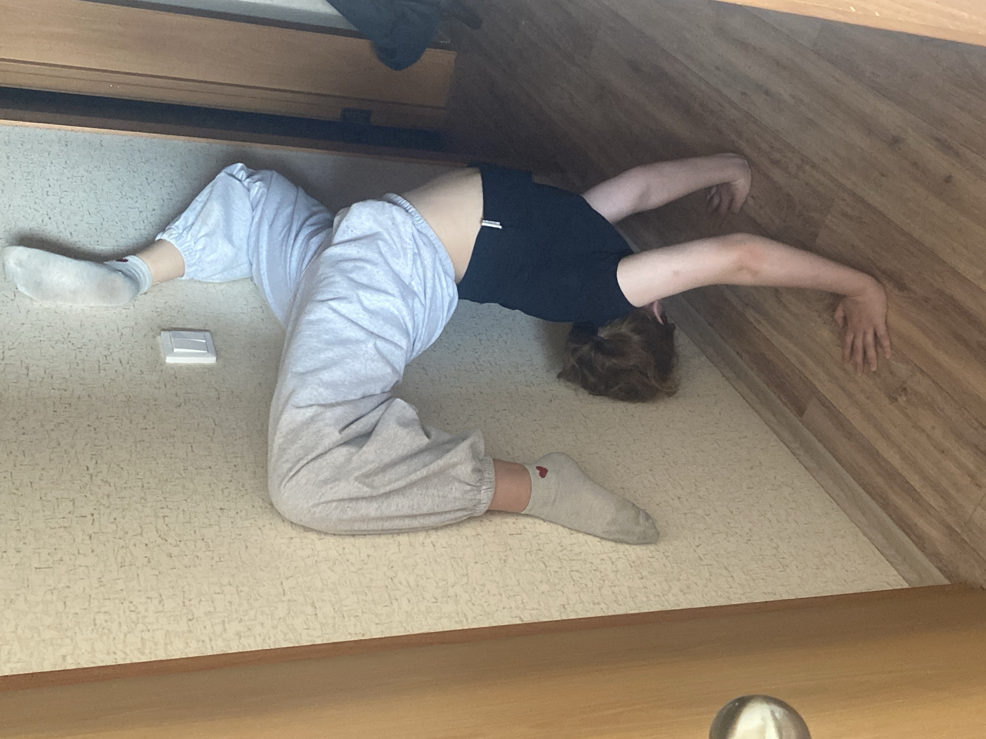 The image captures a person lying on their back on a light-colored, patterned floor indoors, likely in Krasnodar, Russia. The individual, appearing to be young, is dressed in light grey sweatpants and a black t-shirt. Their brown hair is visible against the floor, with their head turned to the left.
The person's body is in a contorted yet seemingly relaxed pose; their legs are bent at the knees with feet on the floor, and their arms are extended above their head and to the sides, resting against a wooden surface that forms part of the wall or furniture. They are wearing light-colored socks; the one on their right foot has a small red heart embroidered on it. A small, white rectangular object rests on the floor near their left foot.
The setting includes a prominent wooden structure, possibly a bed frame or part of a wardrobe, running along the top and right edges of the frame. The floor has a subtle, intricate pattern. The lighting appears natural and bright, suggesting the photograph was taken during the daytime. The scene conveys a candid moment of stretching, playful posing, or unconventional relaxation.