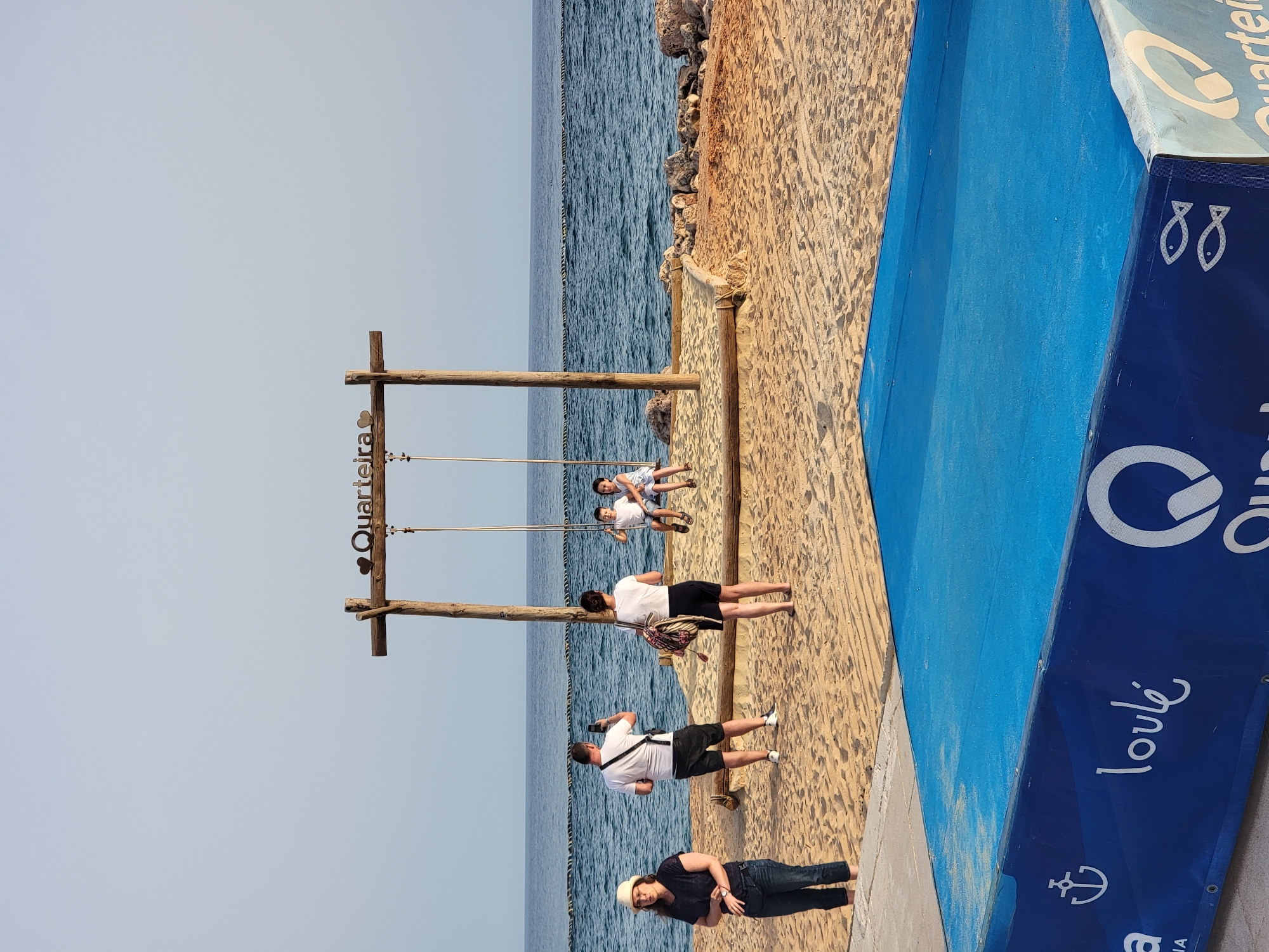 In Quarteira, Portugal, an outdoor scene unfolds on a sandy beach near the ocean. Dominating the frame is a wooden swing set with two children, a boy and a girl, sitting on it. The swing is supported by two sturdy wooden posts, and a wooden beam connects them at the top, inscribed with "Quarteira" and a heart symbol. To the left of the swing, a man stands taking a photograph with his phone, while a woman stands facing away from the camera, holding a bag over her shoulder. Further to the left, a woman wearing a straw hat and jeans stands with her hands clasped. The background features the calm, blue ocean with a thin rope barrier in the water. In the foreground, a vibrant blue rectangular structure is visible, adorned with white graphics, including an anchor symbol, the word "loulé," and a stylized "Q." The sand is textured with footprints and tire tracks, indicating recent activity. The lighting suggests it is late afternoon or early evening.