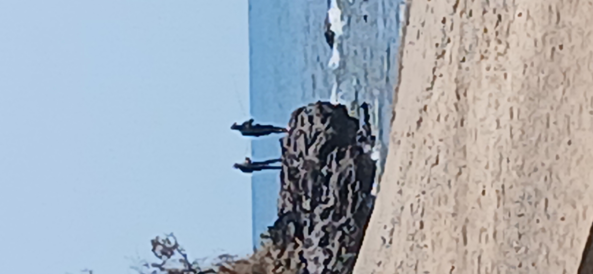 The image depicts two individuals fishing on a rocky outcrop near the coast of Banyuglugur, Indonesia. The scene includes a sandy beach in the foreground, with the ocean visible in the background. The two figures, silhouetted against the light, are standing on a large cluster of dark rocks. Both are holding fishing rods, suggesting they are actively engaged in fishing. The water appears calm with gentle waves lapping against the shore and rocks. The sky is a clear, light blue. The overall impression is one of a tranquil, outdoor recreational activity.