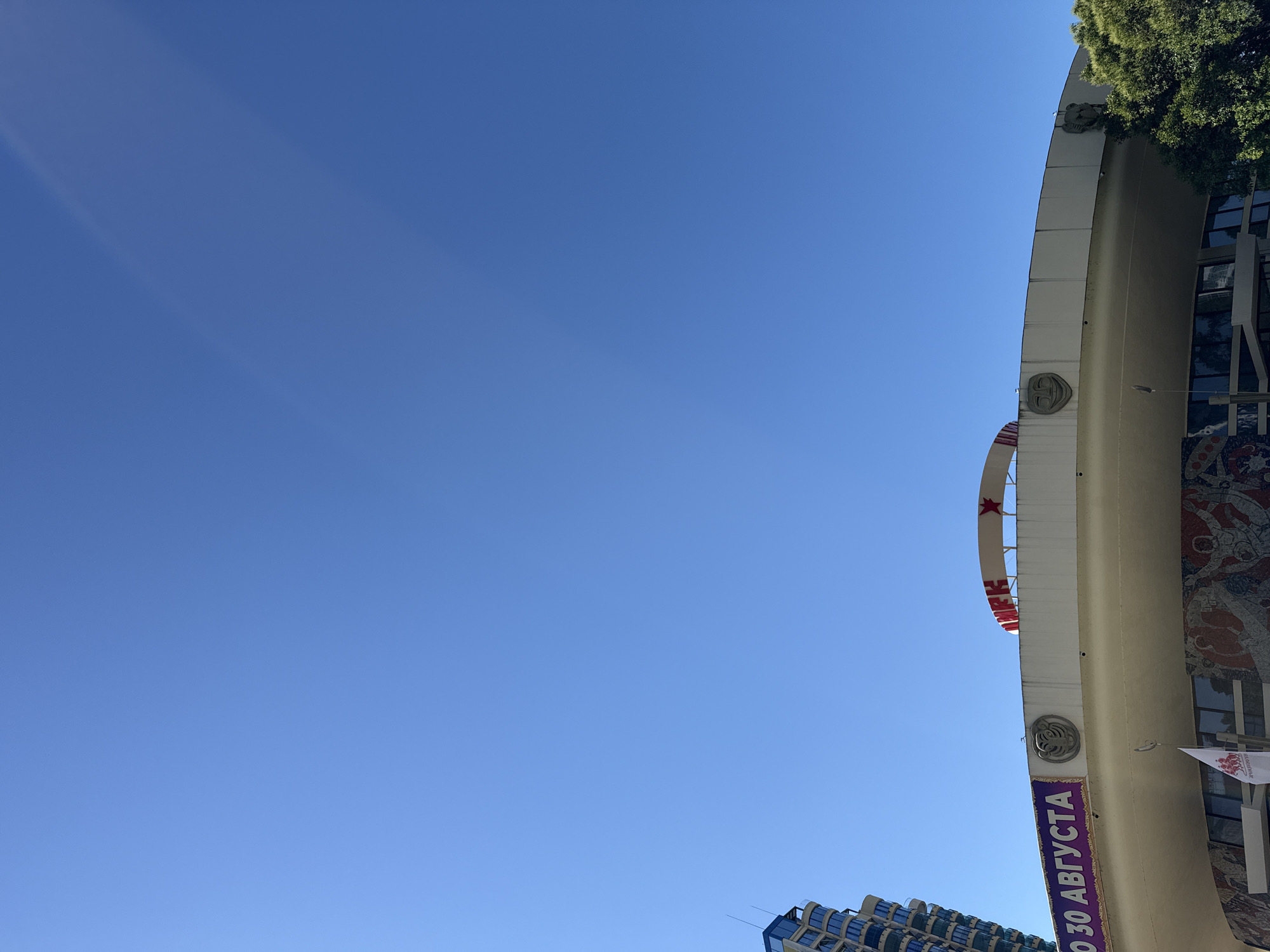 The image captures a bright, sunny day in Sochi, Russia, with a clear blue sky dominating the frame. The perspective is looking upwards at an arched building, likely a cultural or entertainment venue, adorned with architectural details and a large mosaic. Atop the arch, a red star is visible, and lettering in a circular pattern suggests a decorative element. Below this, a purple banner with white Cyrillic text is prominently displayed. The text reads "ДО 30 АВГУСТА," meaning "Until August 30th." The building's facade is a pale cream color, and to the right, a portion of a stained-glass-like mosaic is visible, depicting stylized figures. Trees with lush green foliage are seen at the top of the building, casting shadows. In the background, a modern, multi-story building with a distinctive wavy facade is partially visible. The overall impression is one of a warm, summery day in an urban setting, possibly leading up to a specific event or closing date. There are no people visible in the image.