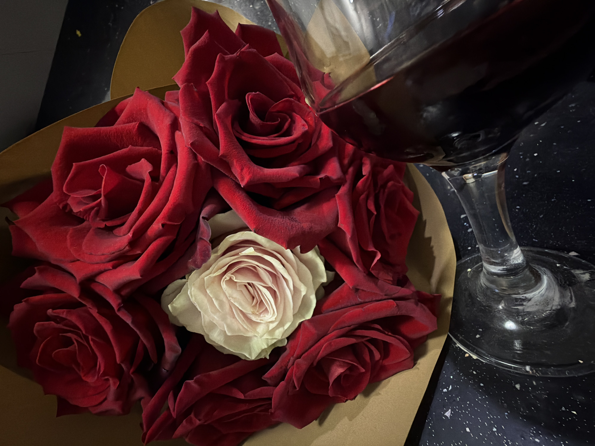 The image displays a close-up, dimly lit composition featuring a bouquet of roses and a glass of red wine. In the center, a bouquet of roses is arranged, predominantly deep red, with one distinct white or very light pink rose positioned centrally among them. The roses are nestled within a light brown, possibly heart-shaped, paper wrapping.
To the upper right, a clear wine glass holds a dark red liquid, likely red wine, with light reflecting off its surface. The base and stem of the glass are visible, resting on a dark, speckled surface, possibly a countertop or table, which is dark grey or black with small white flecks.
The scene suggests an intimate or celebratory moment, potentially an evening or night setting due to the subdued lighting. The focus is entirely on the objects, conveying a mood of romance or appreciation. No people are visible. There is no visible text in the image. This indoor scene is located in Pazardzhik, Bulgaria.