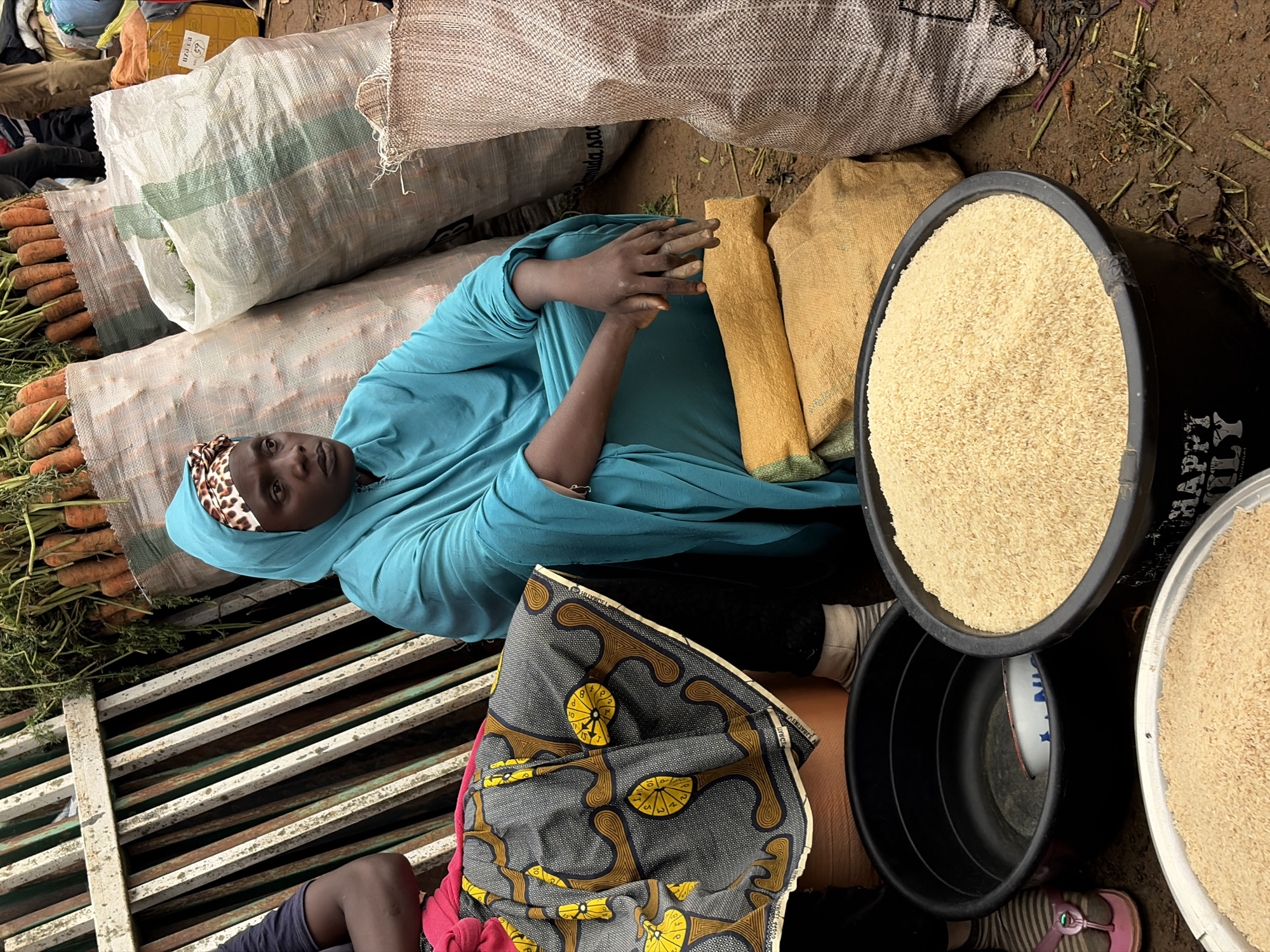 A person wearing a blue headscarf and garment lies among sacks of produce and grain at a market in Gura Top, Nigeria. In the foreground, a pile of carrots is visible, some still with green tops attached, lying next to large woven sacks. To the right, two large black tubs are filled with uncooked rice. A smaller tub is visible inside one of the larger tubs. A patterned cloth is draped over the person's lap. The ground appears to be dirt and littered with debris. The scene suggests a market setting, possibly early morning or late afternoon given the lighting. The person appears to be resting. Text is visible on one of the sacks, appearing to say "CHAMPIONLY", and on a yellow package with text in a foreign language.