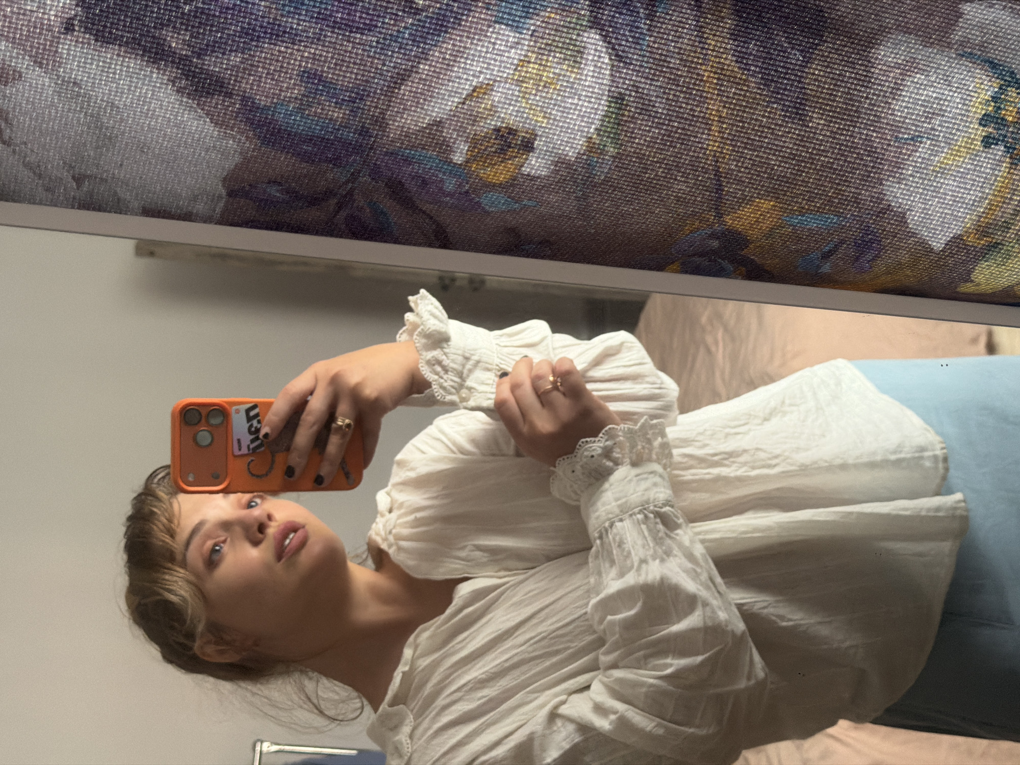 A young woman is captured taking a mirror selfie in an indoor setting. The image is oriented sideways, with the woman's head tilted towards the upper left. She has light brown, wavy hair and light-colored eyes, looking directly into the camera's reflection with a neutral expression and slightly parted lips.
She is wearing a loose, crinkled white blouse with distinct ruffled lace cuffs. On her fingers, she wears two rings, one on each hand. She holds an orange smartphone, likely an iPhone, in a matching orange case. Visible on the phone case is the text "iuen". Her fingernails are painted dark.
The background, reflected in the mirror, shows a patterned textile or fabric at the top, possibly a curtain or tapestry, displaying floral or abstract motifs in shades of purple, blue, yellow, and brown. Below this, a light-colored wall or surface is visible. Behind the woman's body, soft fabrics in light blue and beige tones are present, suggesting bedding or other furnishings. The lighting appears soft and diffused, characteristic of an indoor environment.