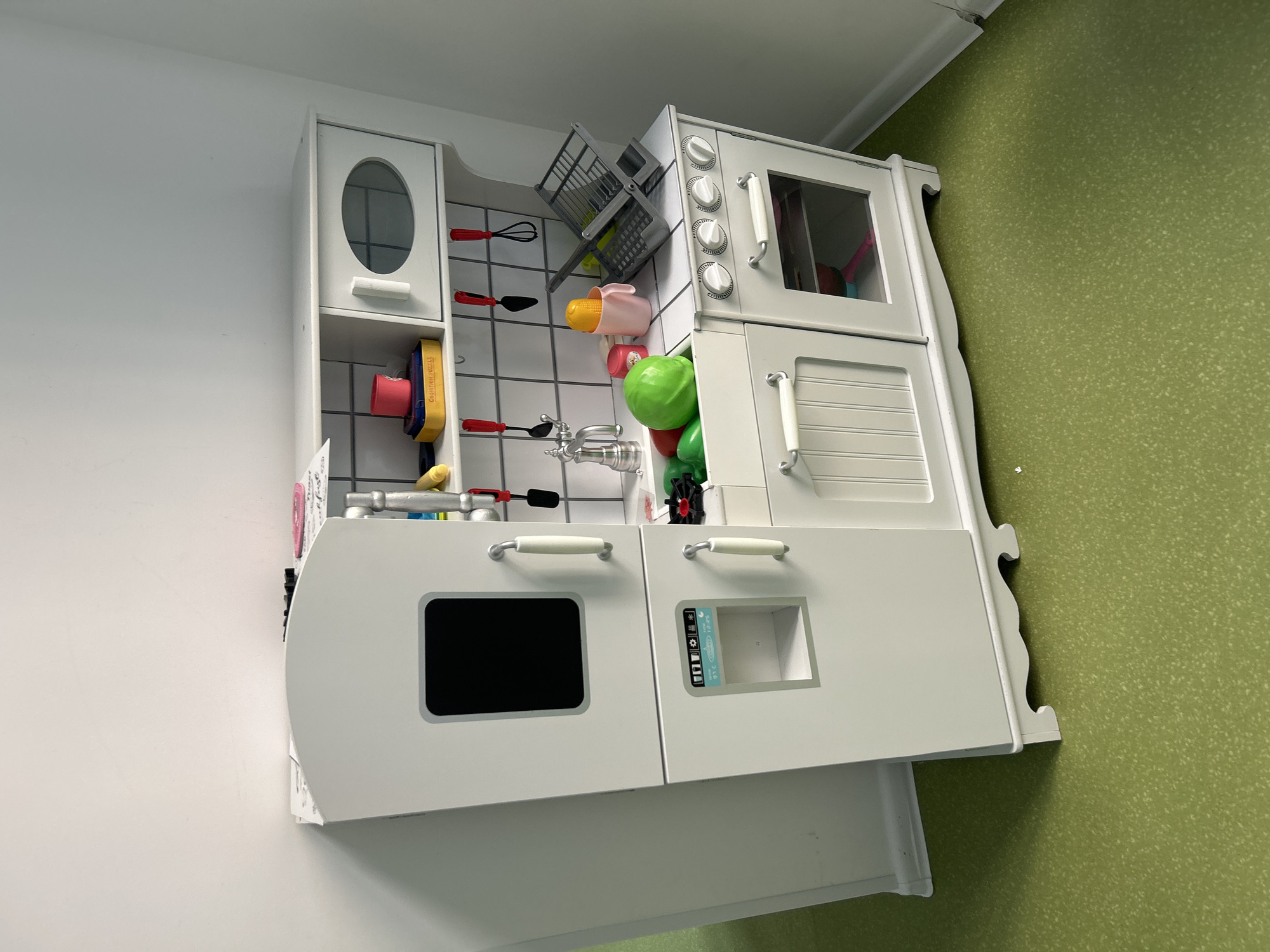 An indoor, eye-level, medium shot captures a white, wooden toy kitchen set against a light-colored wall and a green speckled floor. The kitchen features a refrigerator on the left with a black rectangular screen, followed by a stovetop with multiple knobs and an oven with a glass door. Above the stovetop, there is a sink with a faucet and a tiled backsplash. Shelves are visible above the sink area, holding various toy kitchen accessories such as cups, a yellow corn cob toy, a pink cup, and a green plastic bowl filled with toy produce. Utensils like spatulas and whisks hang from hooks on the backsplash. The overall impression is of a child's play area.
