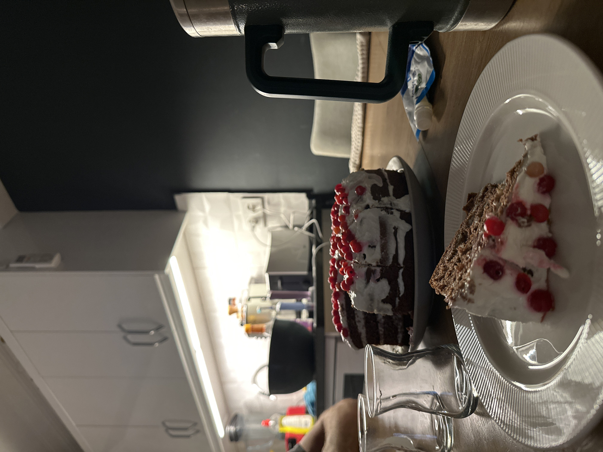 The image depicts a partially eaten chocolate cake decorated with red berries and white frosting, served on a white plate. A slice of the cake has been removed and placed next to the remaining portion. A glass is positioned near the plate, appearing as if it may have been recently used. The scene is set on a wooden surface, possibly a table. In the background, there are kitchen cabinets and a wall. A thermos with a handle is also visible. The location is presumed to be a private residence in the city of Fethiye, Turkey.