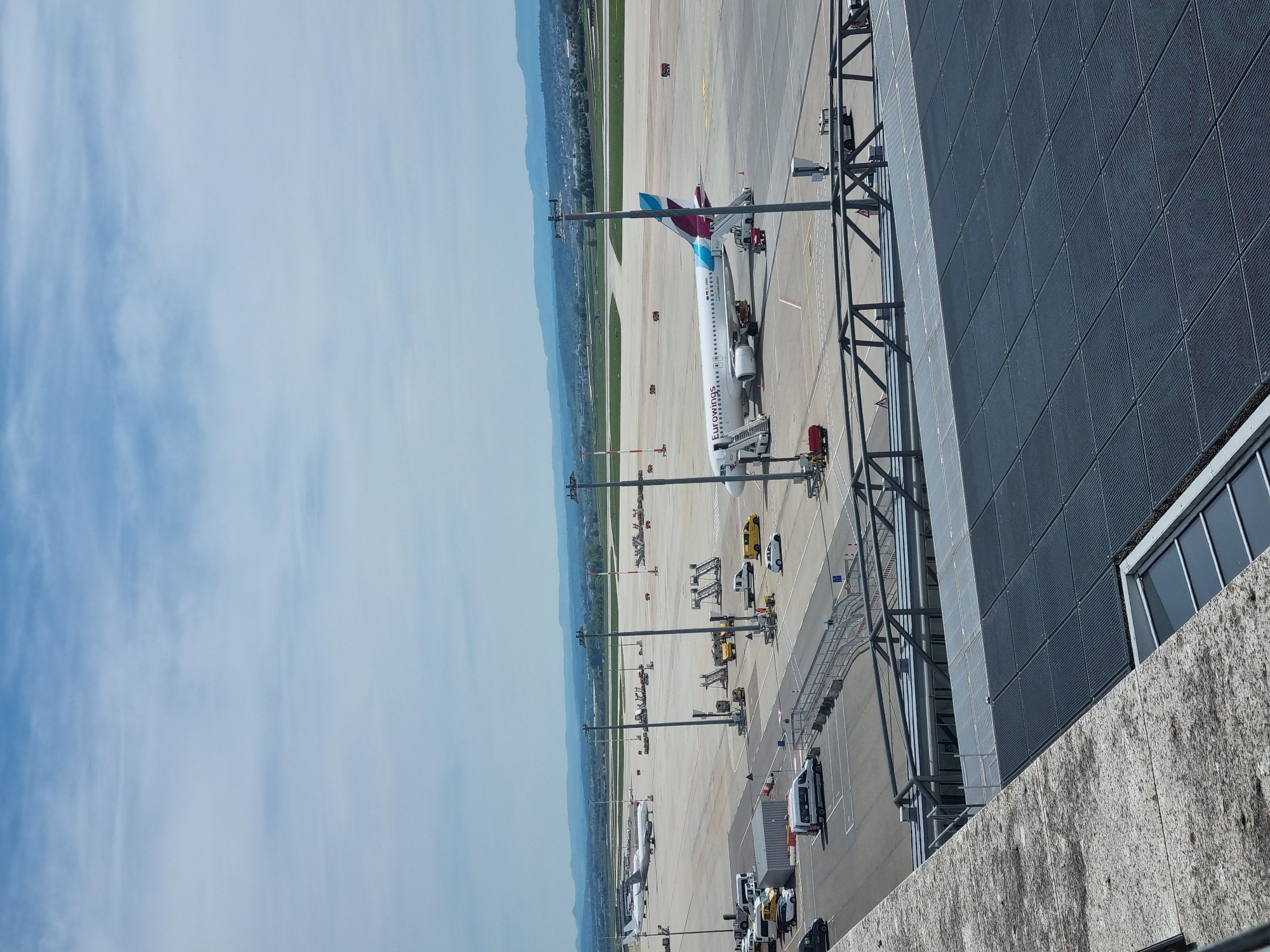 The image shows an airport scene in Echterdingen, Germany. An Eurowings airplane, identifiable by its teal and purple tail, is parked at a gate. A jet bridge is connected to the plane. Various service vehicles are positioned around the aircraft on the tarmac. Light poles are visible along the edge of the tarmac, and in the background, a line of trees and distant hills are present. A portion of the airport terminal building is visible on the right side of the image. The sky is bright blue with scattered clouds.