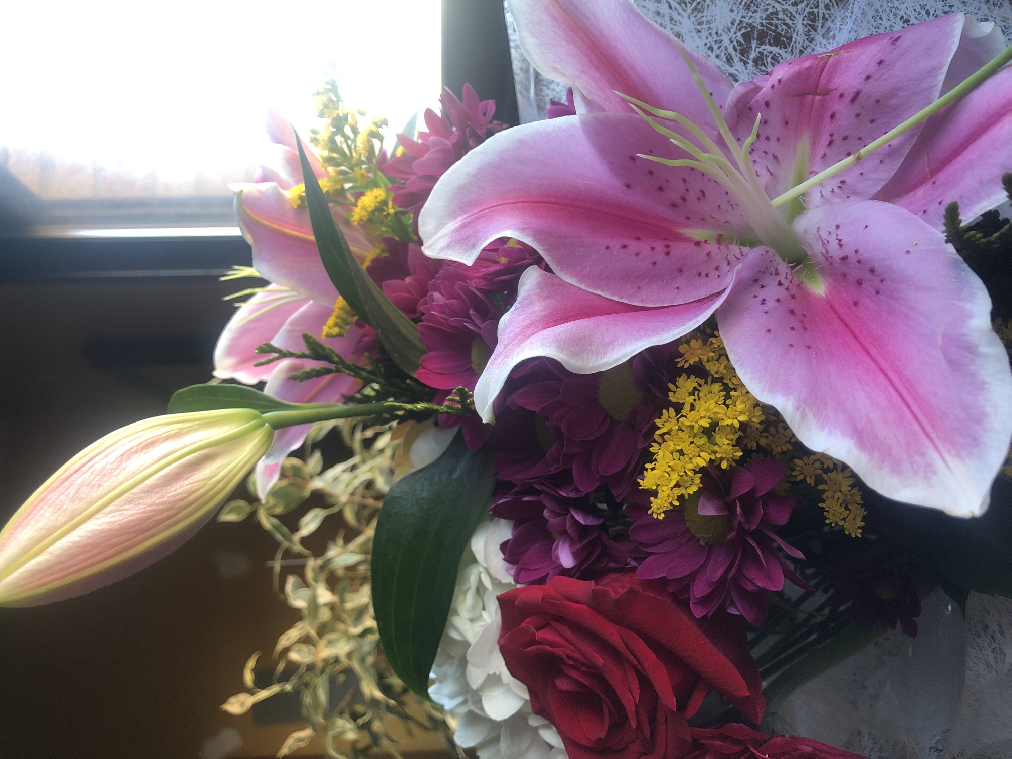 A close-up shot of a bouquet of flowers, including pink lilies with white edges and red and purple chrysanthemums, set against a blurred background. A lily bud is visible on the left. Some white, lacy material is behind the flowers. The location is unknown.