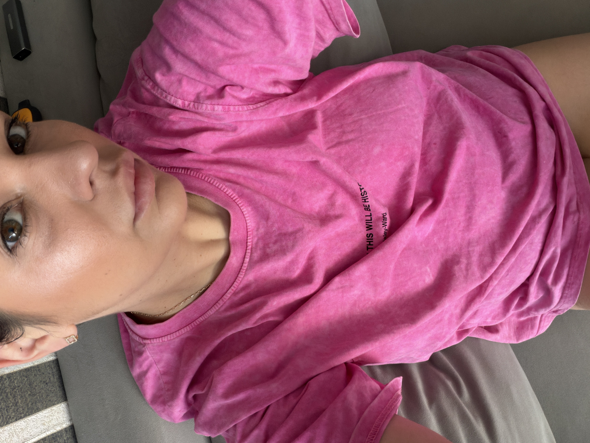 The image shows a close-up, high-angle shot of a woman wearing a pink t-shirt. The t-shirt has black text printed on it. She is lying down on a gray surface, possibly a couch or bed. The background shows striped fabric and a dark rectangular object. The woman's face is partially visible, and she is wearing a small, square-shaped earring. The image does not provide any information about the location in the city of Sochi, Russia.