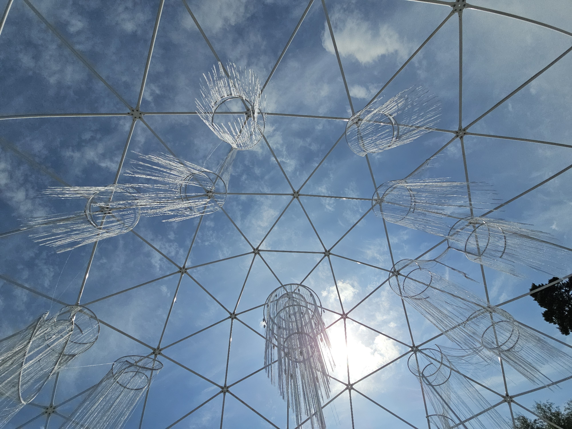 The image shows a view looking up through the geodesic dome structure of the Ulitkino Aviary, located in Ulitkino, Russia. The dome is composed of white metal struts forming triangular panels. Hanging within the dome are several decorative elements that resemble jellyfish or chandeliers. These decorations consist of circular frames from which numerous thin, transparent strands hang down, creating a cascading effect. The sky, a clear blue with scattered white clouds, is visible through the dome's panels. The sun is also visible, shining brightly and causing lens flare. In the lower portion of the image, a dark green tree is visible outside the dome structure.
