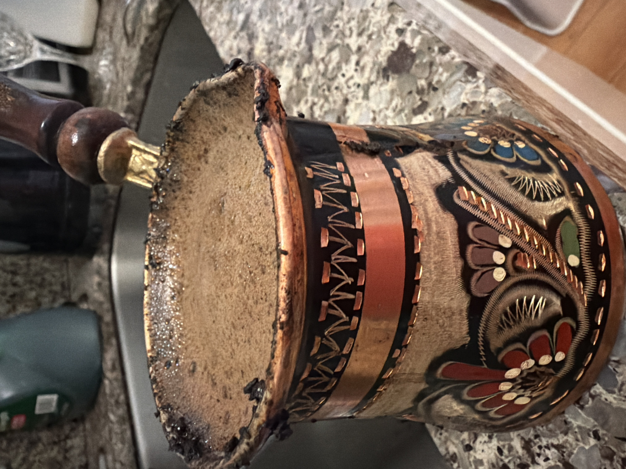A close-up shot shows a copper cezve filled with hot coffee, with some spilled over the rim, sitting on a granite countertop. The cezve has a dark wooden handle with a brass fitting. The countertop is speckled with brown, gray, and white. In the background, a stainless steel sink is visible, along with a blurred green bottle and a wine glass. The scene appears to be in a kitchen.