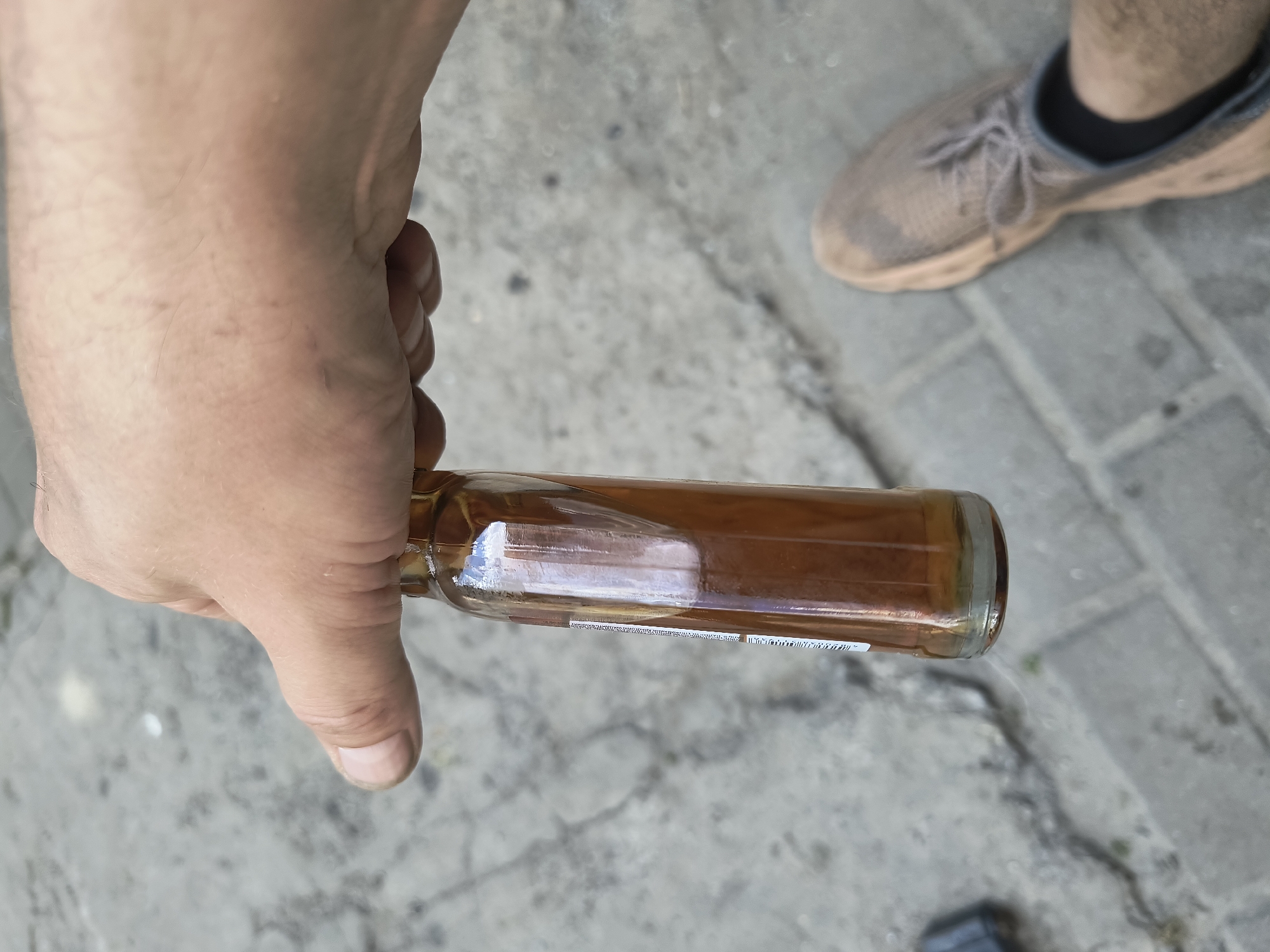 A person's hand is holding a small glass bottle containing a brown liquid. The bottle has a label on the side with text that is mostly illegible. The background is a paved surface with visible cobblestones or pavers. A person's foot wearing a brown sneaker is also visible in the lower right portion of the image. The lighting suggests it is daytime.
