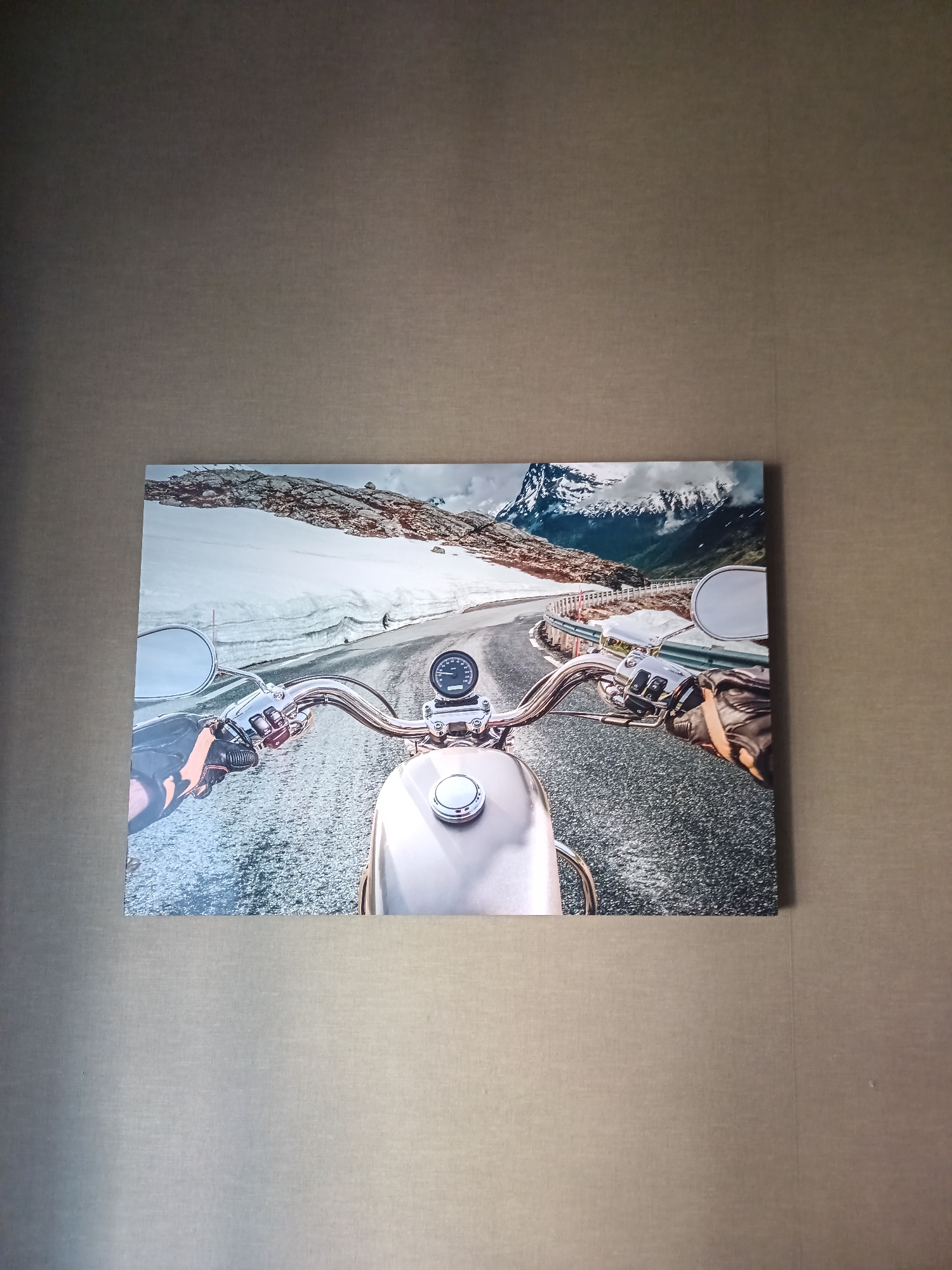The image displays a framed canvas print of a first-person perspective of riding a motorcycle on a paved road with snow-covered mountains and glaciers in the background. The motorcycle's handlebars, speedometer, a chrome fuel cap, and the rider's gloved hands are visible. The print is hung on a textured, grey wall. The location is not specified in the image and cannot be determined.