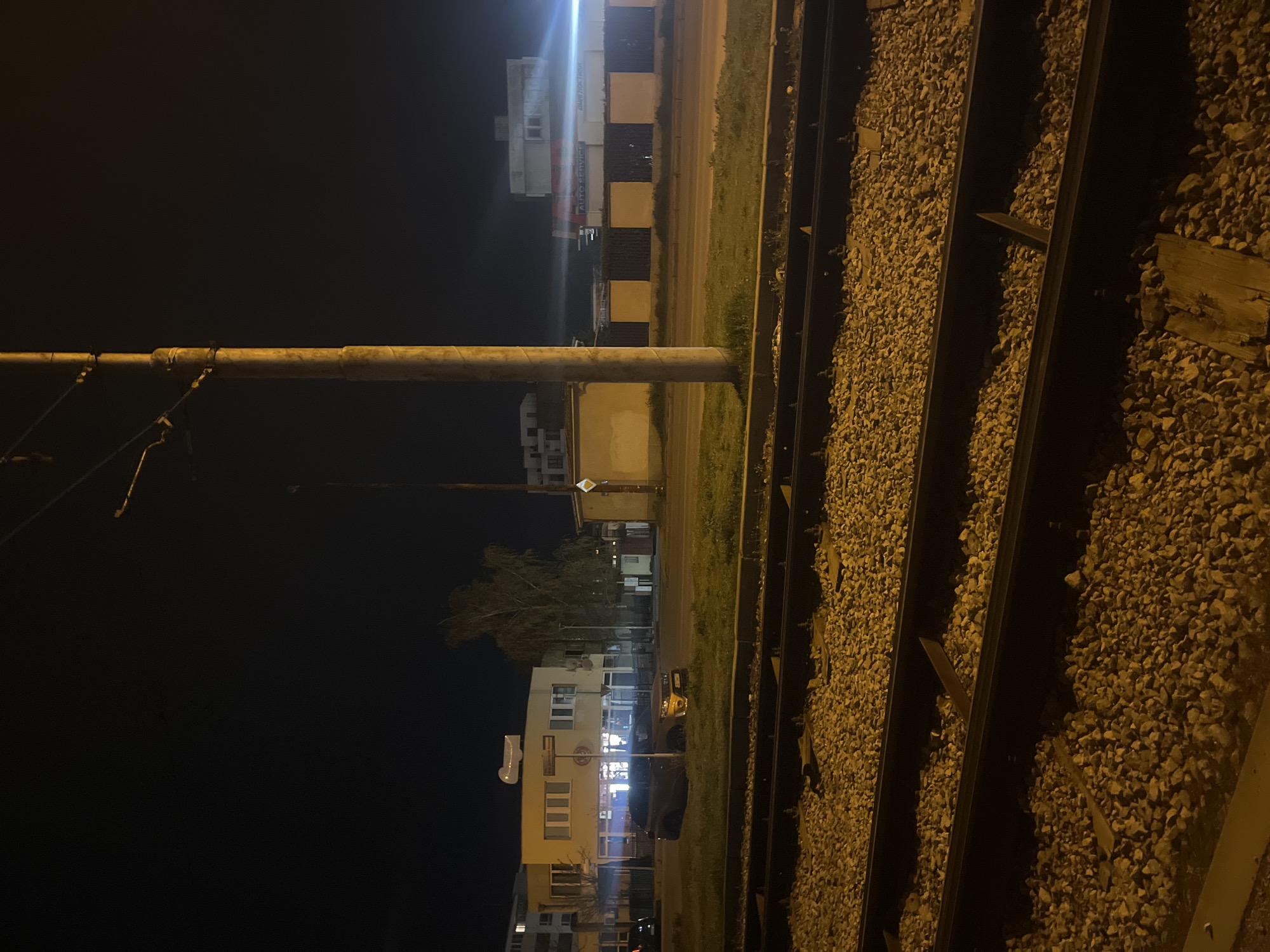 The image shows a street scene in Sofia, Bulgaria, at night. The foreground consists of railway tracks with gravel. A utility pole is visible to the left of the tracks. Beyond the tracks, across a narrow strip of grass and a road, are several buildings. The buildings are illuminated, revealing storefronts and apartments. A car is parked near one of the buildings. The lighting suggests it is nighttime, and the sky appears dark. The scene is viewed from a slightly elevated position.