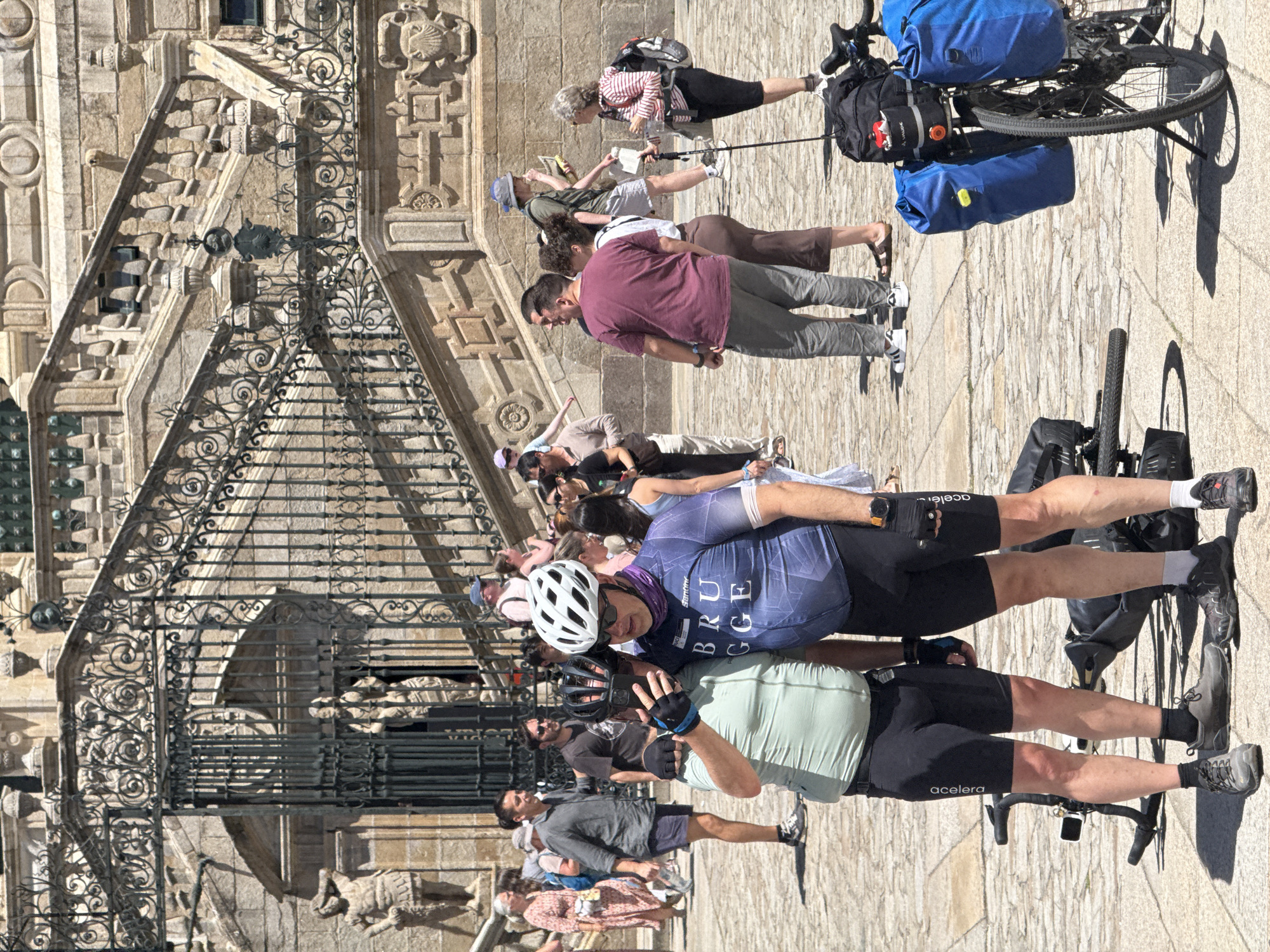 The image shows a sunny outdoor scene in front of a grand stone building with ornate metal gates and a wide staircase. Two men, likely cyclists given their attire and the bicycles visible, are posing for a selfie in the foreground. The man on the left, wearing a light green cycling jersey and black shorts, holds a smartphone and is taking the photo. The man on the right, in a blue and purple cycling jersey with the word "BRUGGE" visible, also wears black cycling shorts and a helmet. Both men are wearing cycling shoes and gloves.
Behind them, other people are milling about, some dressed casually, others appearing to be tourists. The architecture suggests a historical or religious site. The overall atmosphere is that of a public gathering on a bright day. The building's stone facade and the intricate ironwork of the gates and railings point to significant architectural detail. The ground is paved with cobblestones. The presence of bicycles, some with panniers, suggests a touring or recreational context.
The scene is located at the Obradoiro Square in front of the Santiago de Compostela Cathedral in Santiago de Compostela, Spain. The iconic staircase and the Baroque facade of the cathedral are discernible in the background.