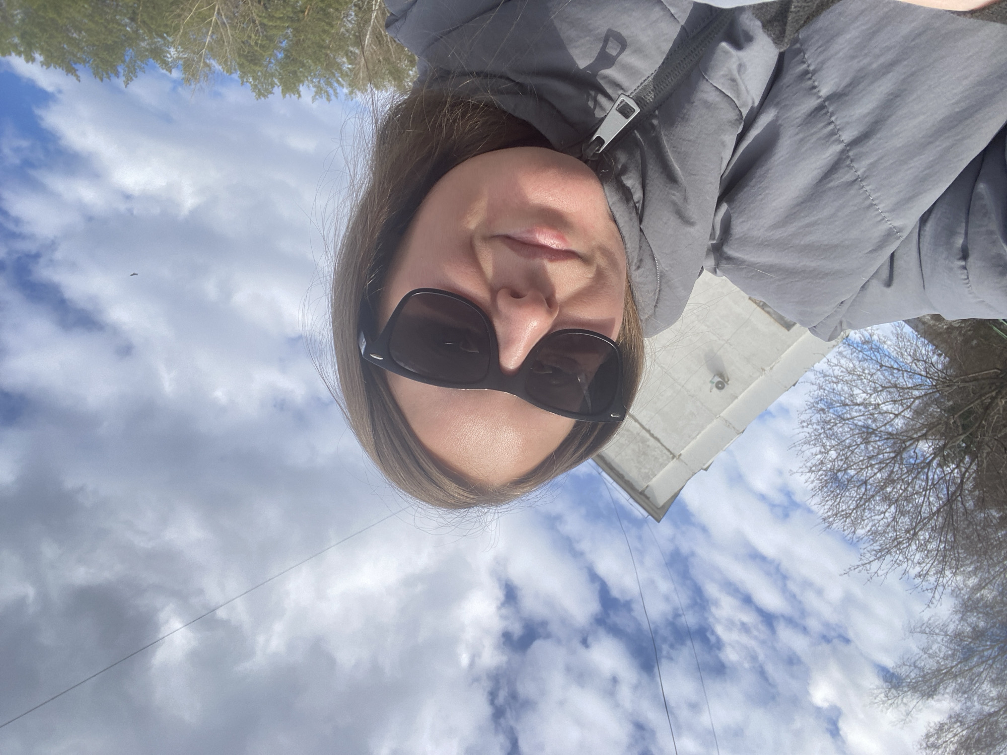 An outdoor selfie captures a woman looking downwards at the camera in Noviy, Russia. She wears dark sunglasses and a grey puffer jacket, with a visible zipper detail near her neck. Her hair is brown and frames her face. The background is dominated by a bright, partly cloudy sky with white and grey clouds.
To the upper left of the image (relative to her face), a dense line of coniferous trees is visible, while to her upper right, bare deciduous tree branches are seen against the sky. A white building structure, possibly a roofline or eave, is partially visible behind her to the right. A thin, dark wire or cable stretches diagonally across the sky in the lower left portion of the background. A very small, dark, elongated object, possibly a drone or distant aircraft, is faintly visible in the sky to the left.
The setting is an open area, likely a park or a natural landscape bordering some human structures. The bare branches of the deciduous trees suggest the season is late winter or early spring. The time of day is daytime, with ample natural light. The woman's expression is obscured by sunglasses, but her lips show a neutral to slight smile. There is no visible text in the image.