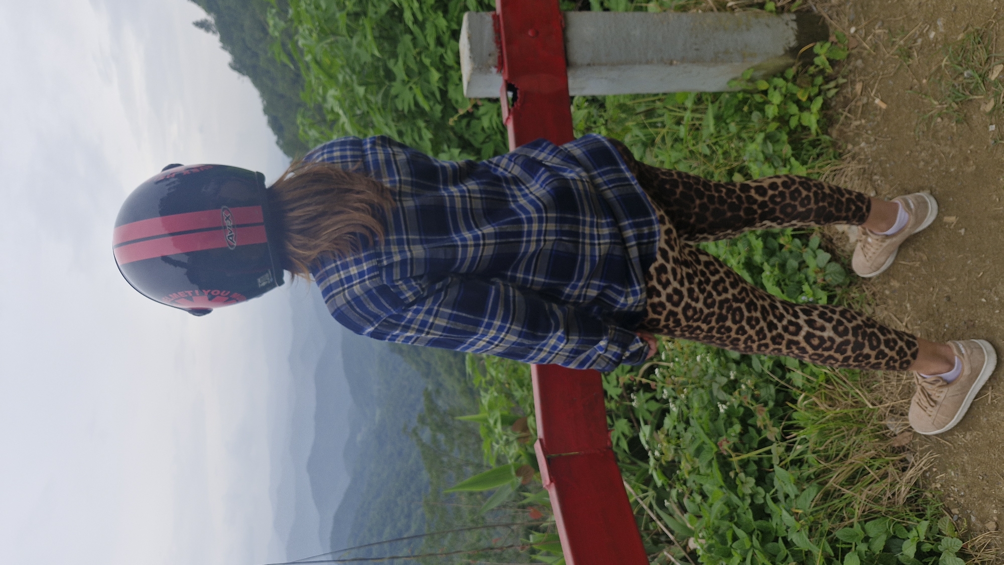 A person stands with their back to the camera, facing a panoramic view of lush, mist-shrouded mountains. They are wearing a dark blue and yellow plaid shirt, leopard-print leggings, and tan sneakers. On their head, they wear a black helmet with two red stripes and the word "AVEX" printed on it. A red railing, partially obscured by greenery, runs horizontally in the foreground. The ground is a mix of dirt and grass. The location is identified as Samoeng Tai, Thailand, suggesting a scenic mountainous area.
