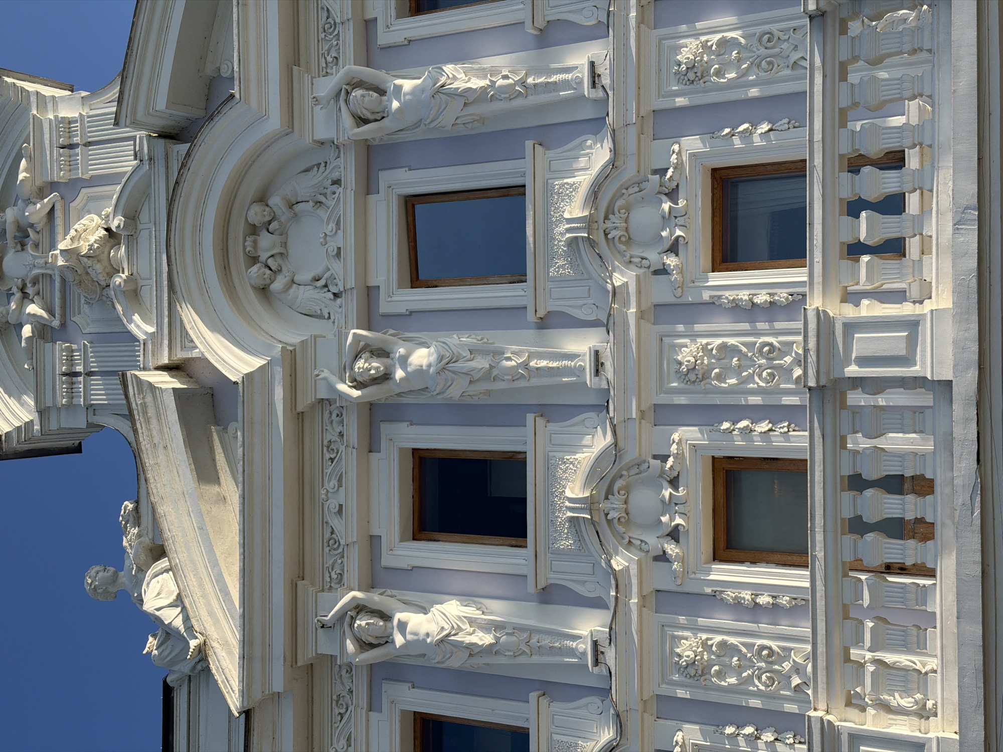 This image shows the facade of a building, likely a historic one, with ornate architectural details. The building's exterior is a light purple color, with extensive white stucco or plaster ornamentation. Key features include:
* **Statuary:** Several female figures are depicted in stucco, acting as caryatids, supporting decorative elements above them. They are draped in flowing garments and appear to be in a pose of exertion. At the very top, on a ledge, is a seated female figure holding a basket of what appears to be flowers. Additionally, there are stucco figures of cherubic children in an arched niche.
* **Windows:** Rectangular windows with wooden frames are visible. Some are open, revealing a dark interior.
* **Balcony:** A white, ornate balcony railing with intricately carved balusters spans the lower portion of the visible facade.
* **Decorative Motifs:** The facade is covered in detailed relief work, including floral patterns, scrolls, and geometric designs.
* **Sky:** A clear, deep blue sky is visible above the building.
The architectural style suggests it could be from the Art Nouveau or a similar decorative period. While the image itself does not explicitly state the location, based on similar architectural styles and common tourist attractions, this building could be located in Nizhny Novgorod, Russia. For example, the **Lyadov House (House of Lyadov)** at Verkhne-Volzhskaya Embankment, 19, Nizhny Novgorod, features similar architectural elements, including caryatids and elaborate stucco work.