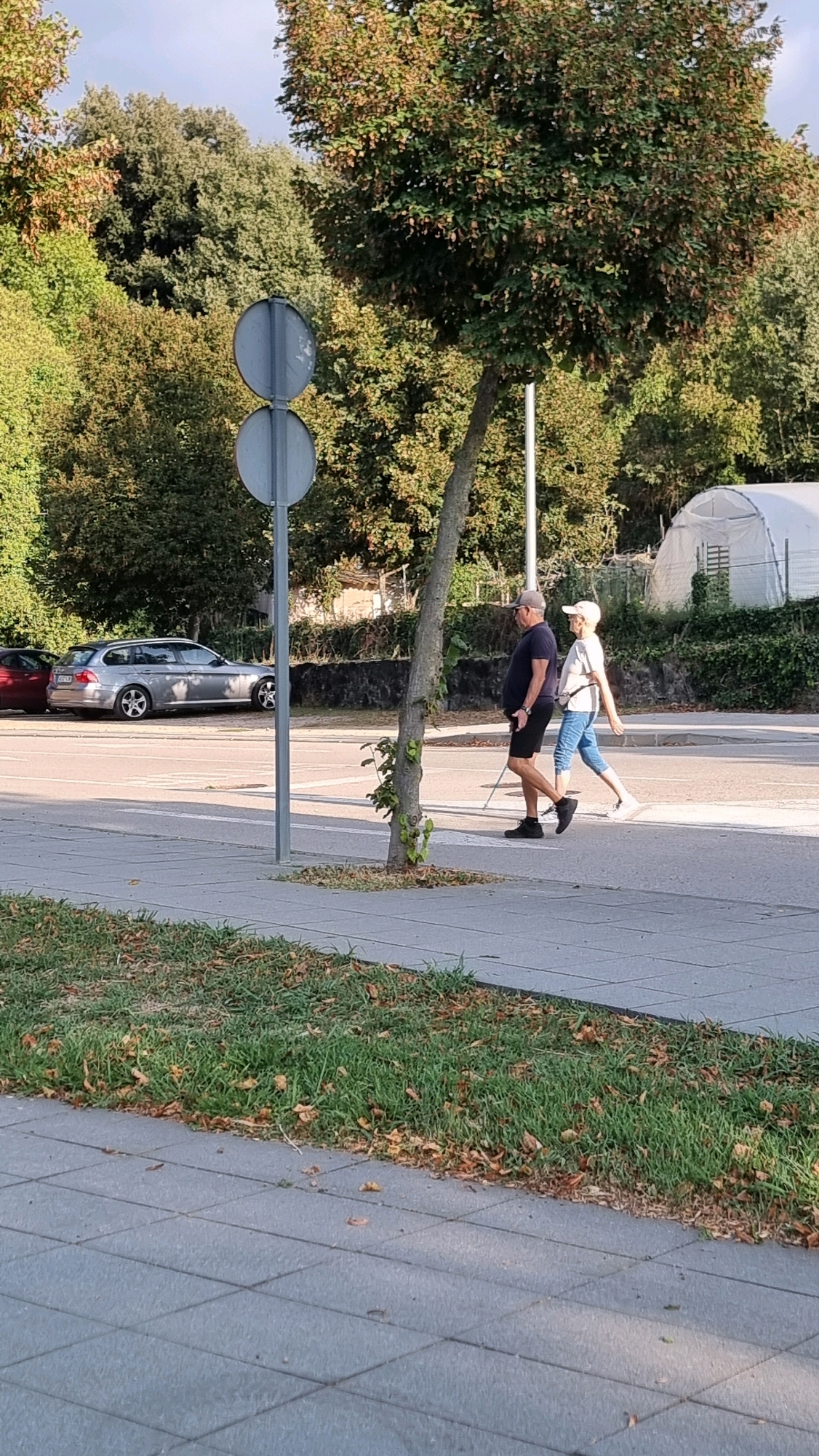 Two people are walking on a sidewalk in front of a road. The person on the left is a man wearing a dark blue t-shirt, black shorts, black shoes, and a grey baseball cap. He is holding a white cane. The person on the right is a woman wearing a white t-shirt, blue jeans, and white sneakers. She has her right arm extended behind her. They are walking from left to right. In the background, there are trees, a silver car, and a white structure that appears to be a greenhouse. The scene is illuminated by natural daylight. The location appears to be Sant Joan les Fonts, Spain.
