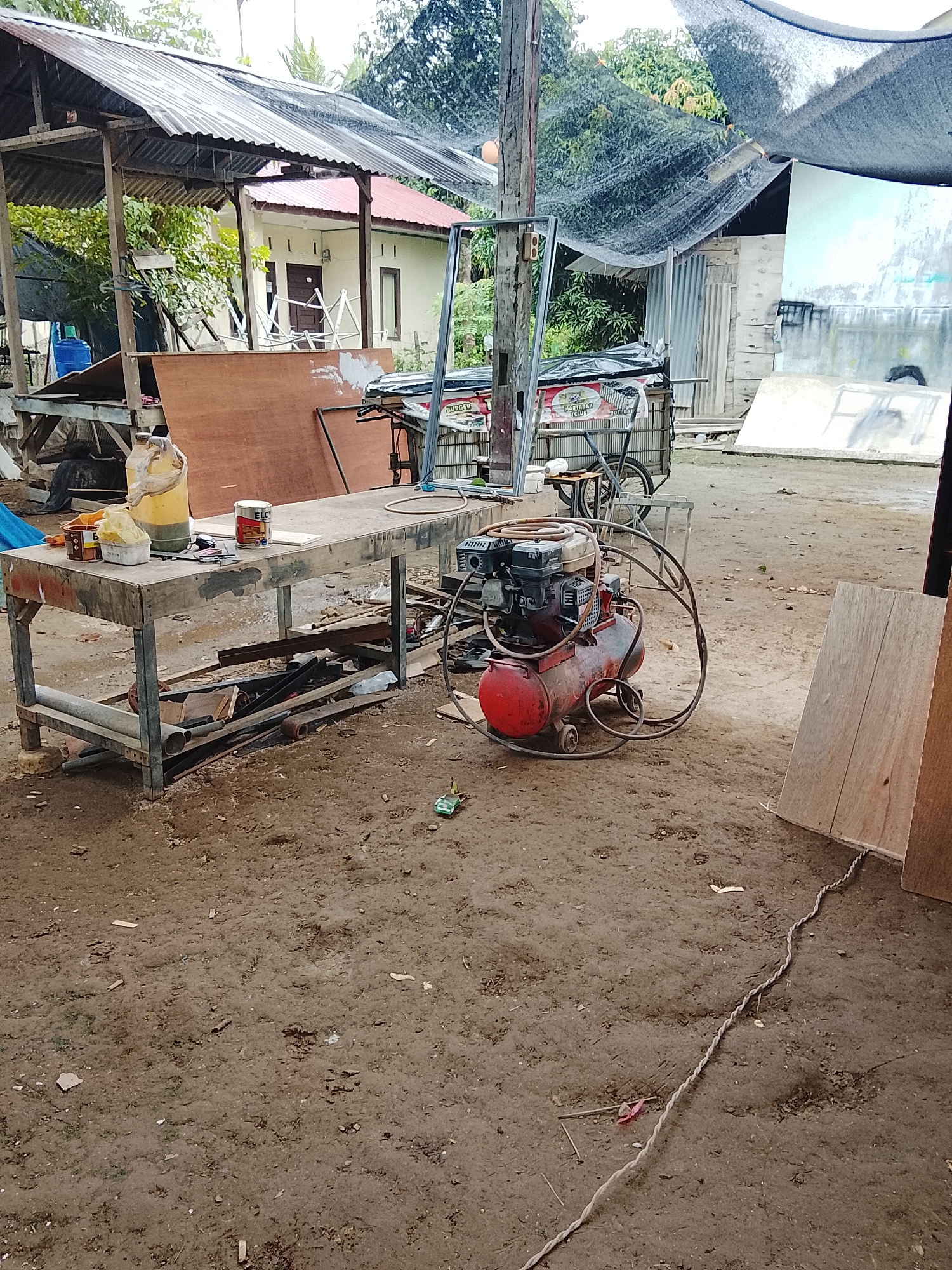 In Mee, Indonesia, an outdoor workshop scene unfolds. The ground is a dusty, unpaved surface scattered with debris, including wood scraps, small pieces of paper, and twigs. In the foreground, a substantial wooden workbench with a metal frame is positioned. On its surface, several items are present: a yellow plastic jug, a red and white can with an "ELOI" label, a small white tub, and various tools. Beneath the workbench, a collection of metal pipes and wood planks are stored.
To the right of the workbench, a red air compressor with a gasoline engine is situated. A coiled black hose is attached to the compressor. Further into the scene, a bicycle is parked near a cart, the cart itself displaying signage with Indonesian text, including "MARTABAK TELUR." Behind the cart, a corrugated metal structure forms a wall.
Overhead, a dark mesh netting is stretched across, providing shade. To the left of the workbench, a simple shelter with a corrugated metal roof supported by wooden beams is visible. Behind this shelter, a white house with a red roof and brown window frames stands, partially obscured by trees. The overall environment appears to be a working area, possibly for construction or repair, with materials and equipment laid out in a functional, albeit somewhat disorganized, manner.
