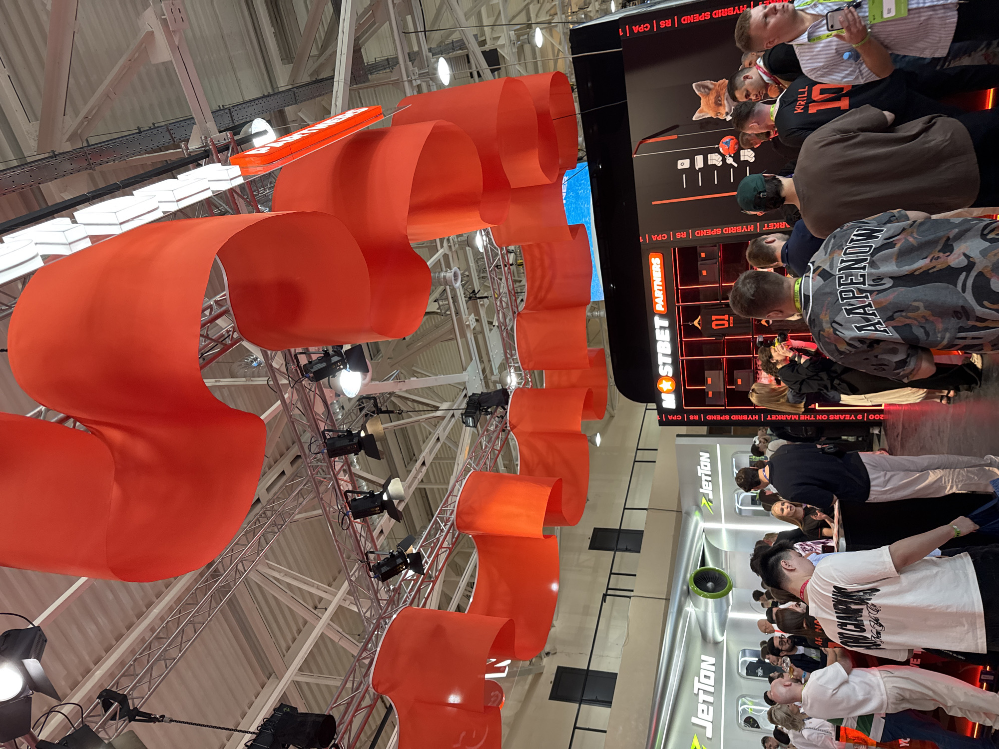 The image shows a crowded indoor space, possibly a convention or trade show, with red banner-like structures suspended from the ceiling. The ceiling is characterized by a grid of metal beams and lighting fixtures. Attendees are seen gathered near exhibit booths. Signage for "JetTon" and "MSTBET PARTNERS" is visible. Text indicates "9 YEARS ON THE MARKET | HYBRID SPEND | RS | CPA | 1". Several people are visible, some wearing name tags, indicating that they are part of the event. The location is Yerevan, Armenia.