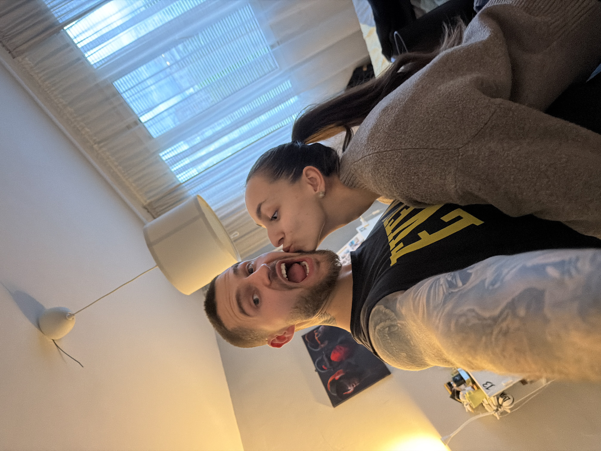 An indoor shot captures a playful moment between a man and a woman, oriented with a significant counter-clockwise rotation, making the scene appear on its side.
The woman, with dark hair pulled back in a ponytail and a small earring, wears a light brown, textured long-sleeved top. She is positioned to the left, leaning in to kiss the man's cheek with her eyes gently closed or cast downwards.
The man, to the right, has short dark hair and a beard. He is making a humorous expression, with his mouth wide open, showcasing his teeth and part of his tongue. He wears a black t-shirt with a yellow design or text on the chest, partially visible. His muscular arms are adorned with extensive tattoos that resemble full sleeves. His eyes are wide, contributing to the comical effect of his facial expression.
In the background, a large window with white horizontal blinds or sheer curtains provides ambient light. Below the window, on the left, a white cylindrical light fixture hangs, and further down, a round white object with a wire attaches to the wall, possibly a pull switch or a component of the lighting system. Behind the man's head, a dark framed artwork featuring abstract red and black shapes is mounted on the wall. To the far right, on a surface, some indistinct objects are visible, including a white item with the number "13" printed on it. The overall scene suggests an affectionate and lighthearted interaction between a couple in a domestic setting, likely during the daytime.