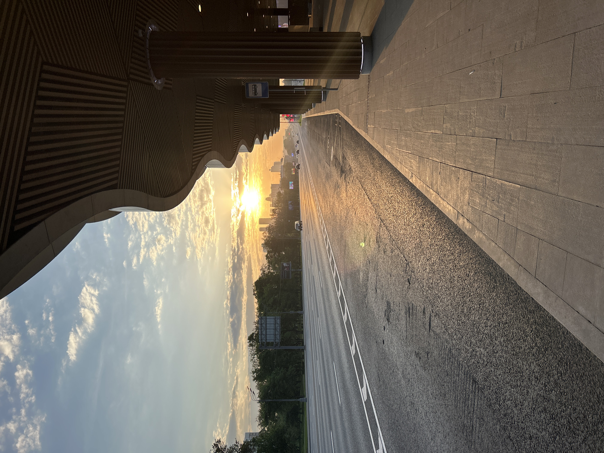 Here is a description of the image:
The image presents a ground-level, urban scene at what appears to be either sunrise or sunset. A multi-lane road stretches away from the viewer, leading toward a city skyline silhouetted by the sun on the horizon. There are some vehicles visible on the road. Along the side of the road, a stone-paved walkway is visible. At the edge of the walkway, there is a structure with a unique, wave-like architectural feature overhead, supported by columns. A small blue bus stop sign is visible along the structure. The sky is blue with scattered white clouds, while the sun casts a warm, golden light across the scene. Trees line the road further down in the distance.