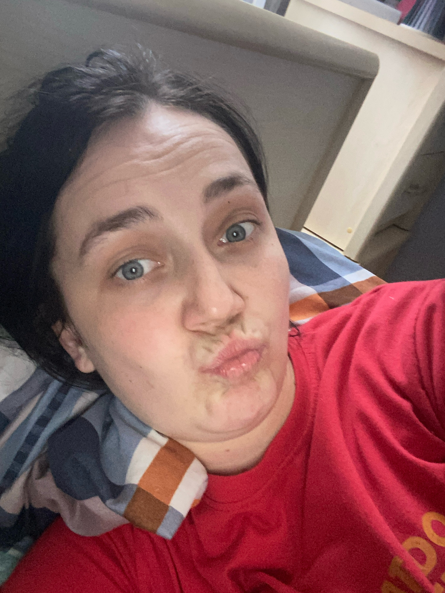 In a selfie taken from a high angle, a woman with dark hair and blue eyes makes a puckered face. She is lying on a bed with a plaid pillow and wearing a red t-shirt. The background consists of a beige headboard and a dark gray wall. The lighting is soft and diffused, suggesting an indoor setting.
