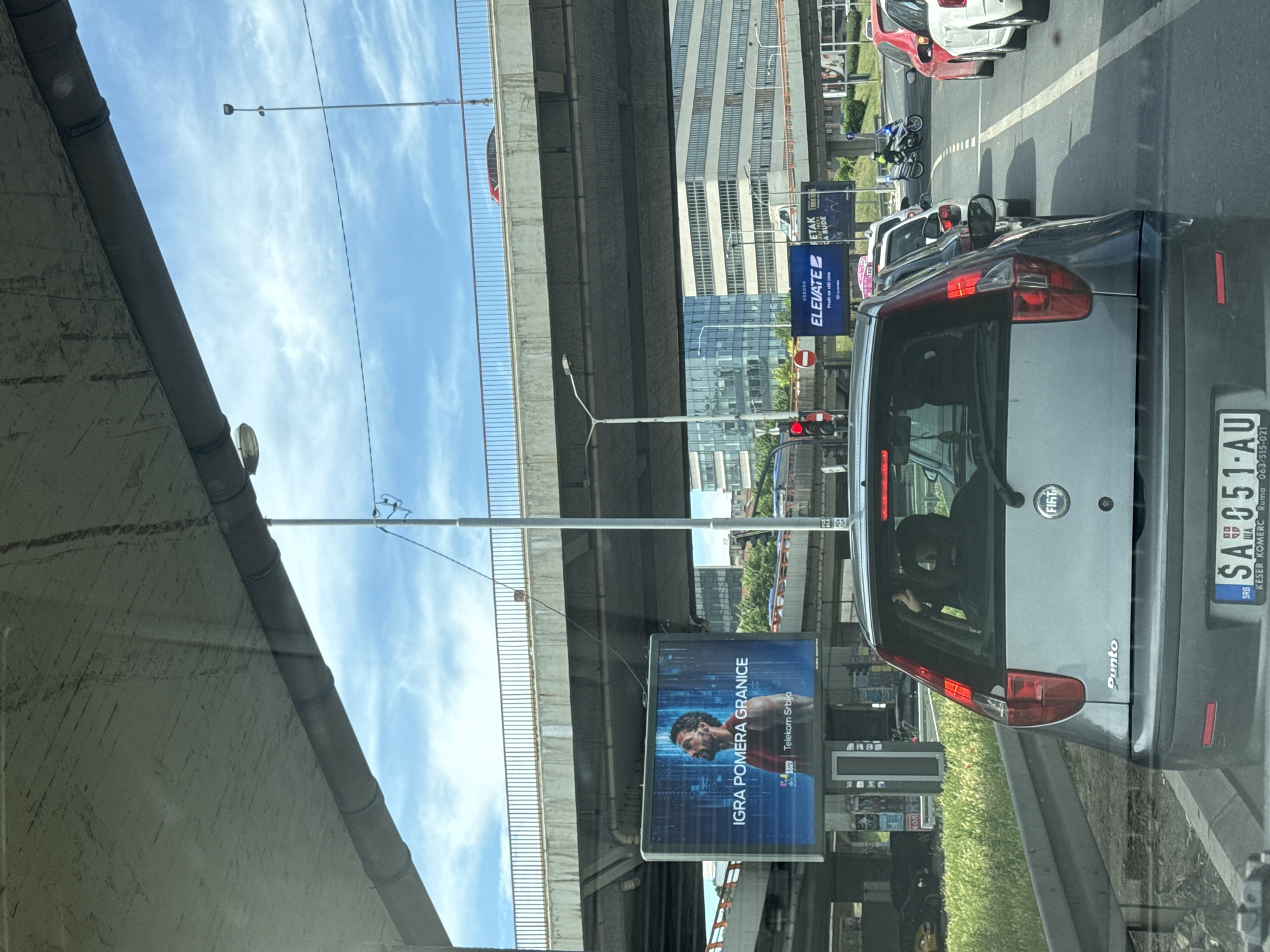 The image captures a daytime street scene, likely from inside a car stopped in traffic, looking forward. The main subjects include several cars, a large concrete overpass structure overhead, and multiple billboards lining the road.
In the foreground, the rear of a grey Fiat Punto car is prominent, displaying a Serbian license plate "SA 051-AU". Below the license plate, text "KESER KOMERC Ruma 063/515-021" is visible. The car has "FIAT" and "Punto" badging. Further down the road, other cars are stopped, including a white and a red vehicle, suggesting a traffic jam. A red traffic light is visible in the mid-ground, confirming the stopped traffic.
The setting appears to be an urban environment, characterized by tall buildings in the background, a multi-lane road, and the significant overpass structure that stretches across the frame. The sky is bright blue with scattered white clouds, indicating clear weather.
Several large advertising billboards are visible. One prominent billboard features a man and the Serbian text "IGRA POMERA GRANICE" (Game moves boundaries) with the logo "Telekom Srbija". Another billboard further back reads "USKORO ELEVATE" (ELEVATE soon) and "Predi na viši nivo" (Move to a higher level). Other partial text like "ETAK" and "DA BUDE" can also be seen on smaller signs or parts of billboards. Streetlights are also part of the urban infrastructure. The concrete walls and pillars of the overpass dominate the upper-left and central parts of the image.