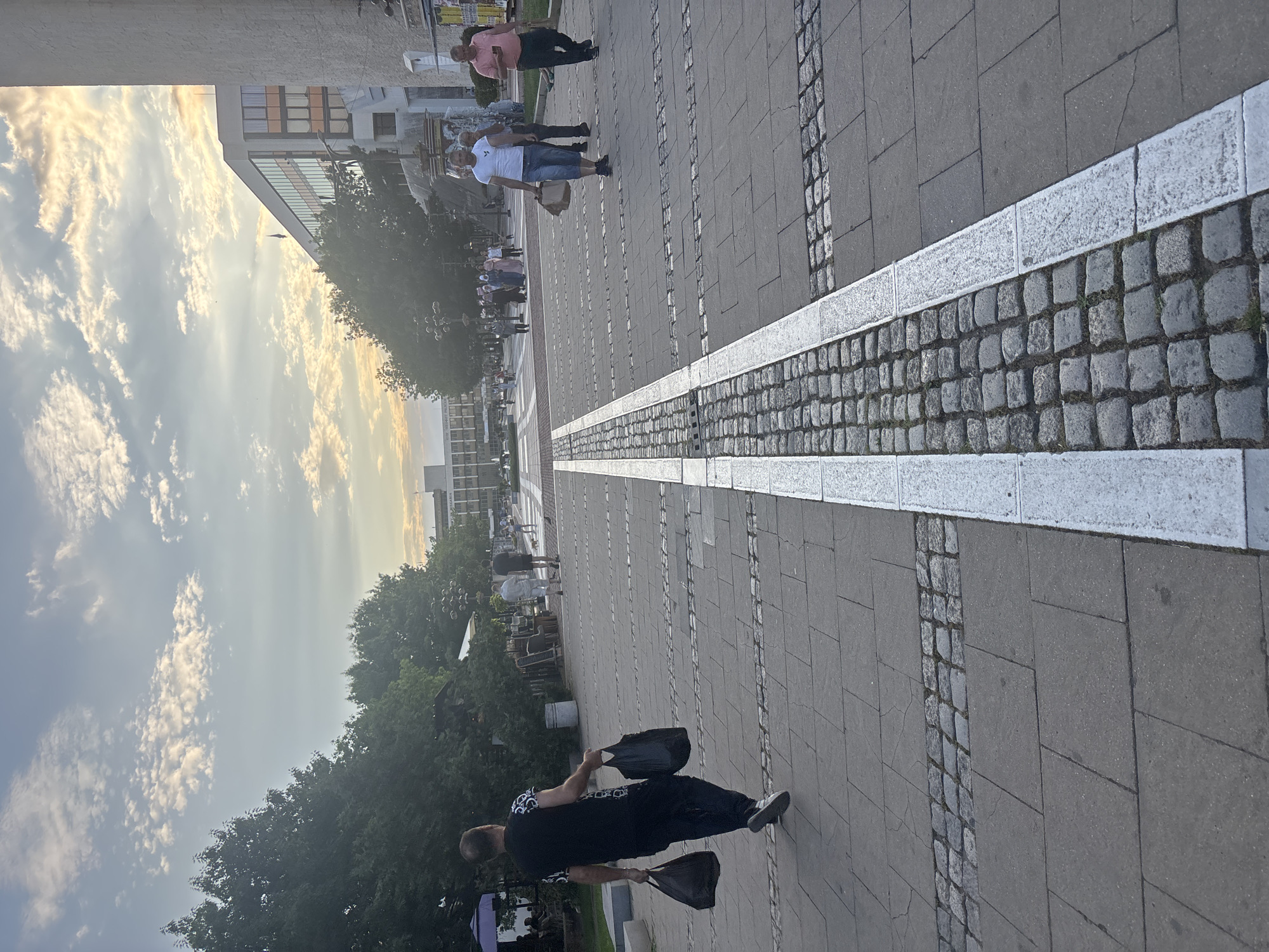This image depicts a wide pedestrian area or plaza in the city of Blagoevgrad, Bulgaria, during late afternoon or early evening, characterized by warm, soft light. The sky exhibits scattered clouds, with some illuminated by the low sun, displaying hues of orange and yellow towards the right horizon.
In the foreground, the ground consists of grey concrete paving slabs interspersed with horizontal bands of darker, square cobblestones. A prominent central strip of grey cobblestones, approximately 1.5 meters wide, runs vertically through the scene, bordered on both sides by white-painted lines or light-colored stone strips.
On the left side of the pedestrian area, a man is walking away from the camera, carrying a dark plastic bag in each hand. He is wearing a black t-shirt with a white pattern on the upper arms/shoulders, dark trousers, and white sneakers. To his left, a dense line of mature green trees borders the plaza, beneath which are visible outdoor seating arrangements, including tables and a purple umbrella. A dark grey public waste bin is situated near the trees.
On the right side of the pedestrian area, closer to the midground, a man in a white t-shirt and blue denim shorts is walking towards the right, holding a brown paper bag in his left hand. Behind him, another man in a dark shirt is partially visible. Further to the right, a man wearing a pink polo shirt and dark trousers walks, looking at a smartphone in his left hand and carrying a light-colored bag in his right. In the deeper midground, numerous pedestrians are present, including individuals wearing traditional head coverings and a child.
Architecturally, a modern building with a multi-story glass facade and light-colored panels is visible in the upper right background. A dark blue flag is flying from its roof. Adjacent to it on the far right is a tall, light-colored, tower-like structure, part of a larger, older building. Further in the distant background, several other buildings with repetitive window patterns are visible. The pedestrian area extends into the background, where more people are observable. Several multi-globe streetlights are scattered throughout the plaza.