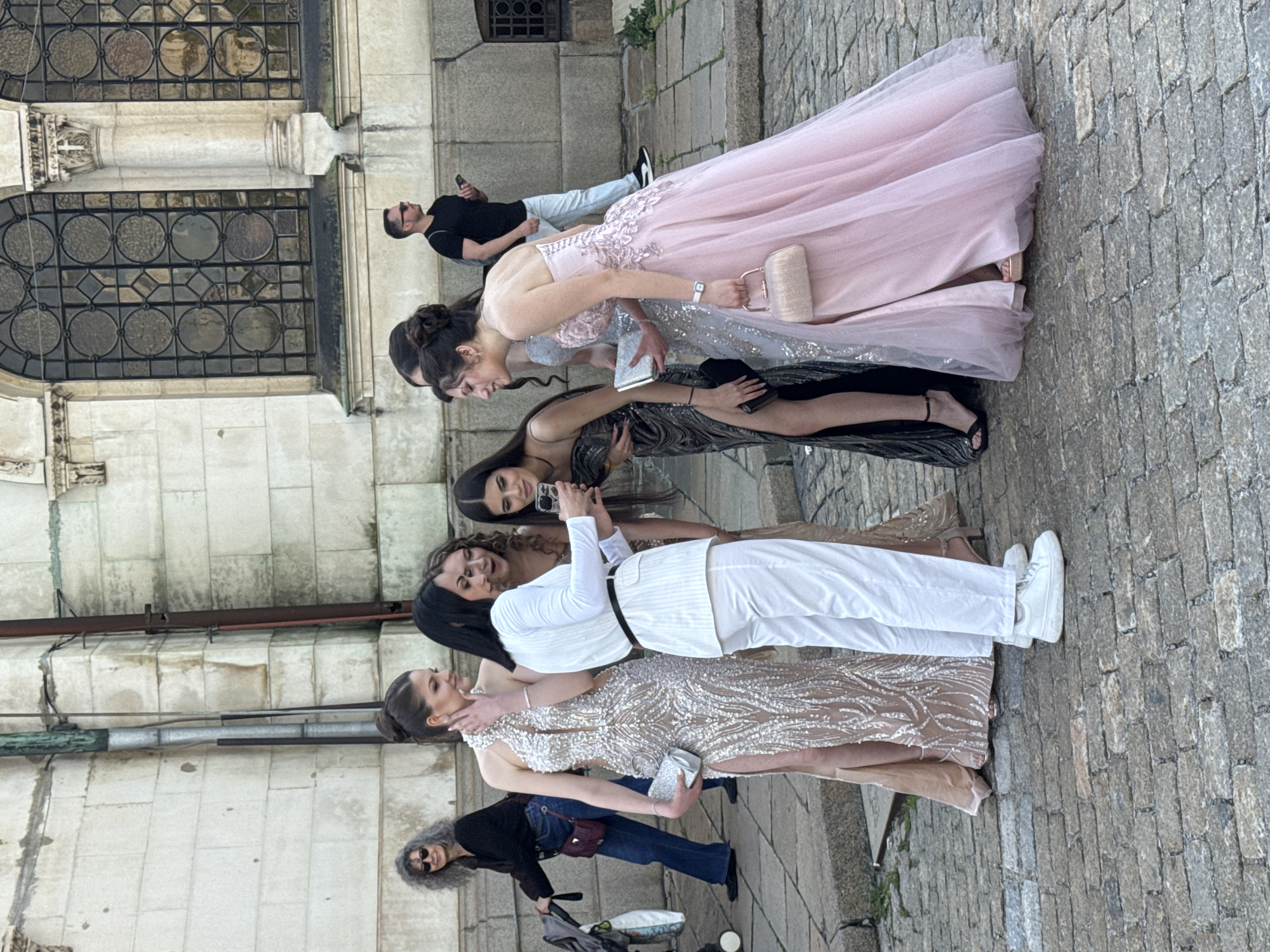 A group of six people, primarily young women in formal attire, are posing for a photo in front of a stone building with large windows. The women are standing closely together, with arms around each other. They are dressed in prom gowns that are pink, silver, black and gold. One person wears a white pants suit. One woman is holding a cell phone, presumably to take the photo. To the left of the group, an older woman stands with a baby stroller. To the right of the group, a man stands holding a cell phone. The setting appears to be outdoors on a stone paved walkway. The architecture of the building in the background suggests an urban environment, possibly in Sofia, Bulgaria. The city of Sofia is not evident, although the architecture of the building and the style of paving suggest a historical section of a city.