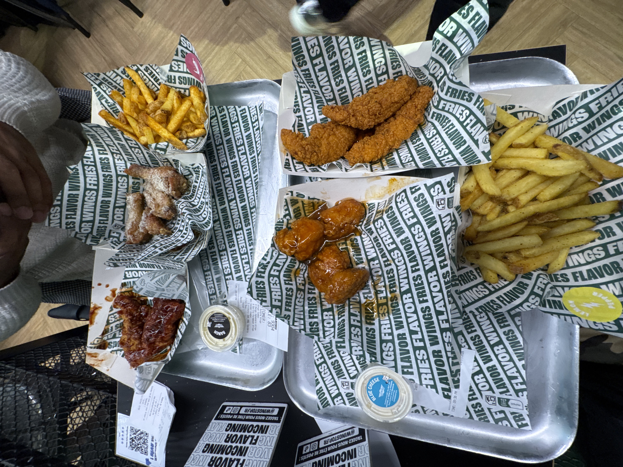The image shows two metal trays laden with food from Wingstop, a fast-food chain specializing in chicken wings. The food includes various flavors of wings and tenders, and fries. The trays are placed on a black, possibly metal, surface. Several take-out containers and receipts are also visible on the trays. Text in French can be seen on stickers placed on the tray. The presence of french text may suggest this image was taken in France.