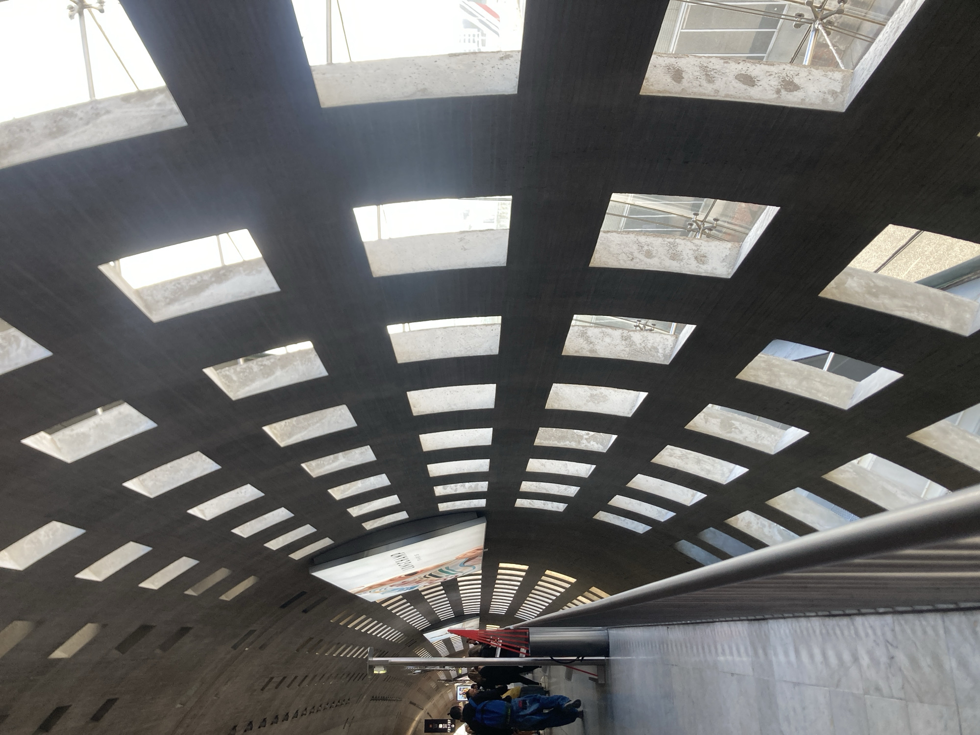 A low-angle shot within a curved tunnel-like structure shows a patterned ceiling with rectangular and square openings allowing natural light to filter through. The ceiling is grey with off-white accents surrounding the openings. A horizontal metal railing runs along the right side of the frame. In the lower part of the tunnel, a small group of people walks towards the vanishing point, and some are visible on a red moving walkway. A billboard is visible on the left. The location is Le Mesnil-Amelot, France.