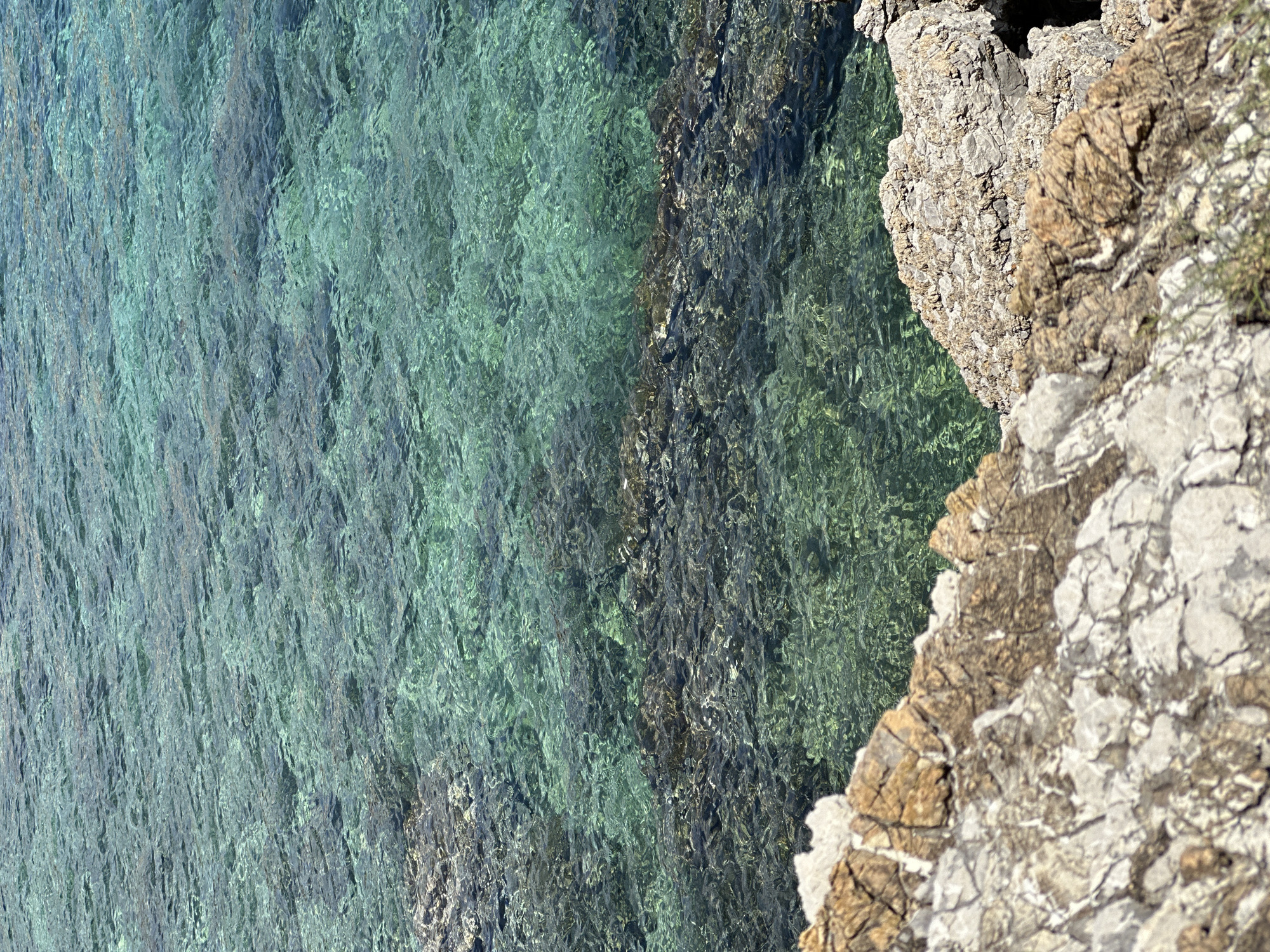 The image depicts a rocky coastline meeting clear turquoise water, likely near Budva, Montenegro. The right side of the frame shows a rugged, textured rock formation with shades of gray, brown, and white. Vegetation is sparse. To the left, the water is transparent, revealing darker rock formations beneath. The water surface is slightly rippled, creating a shimmering effect. The perspective is an overhead view, tilted slightly.