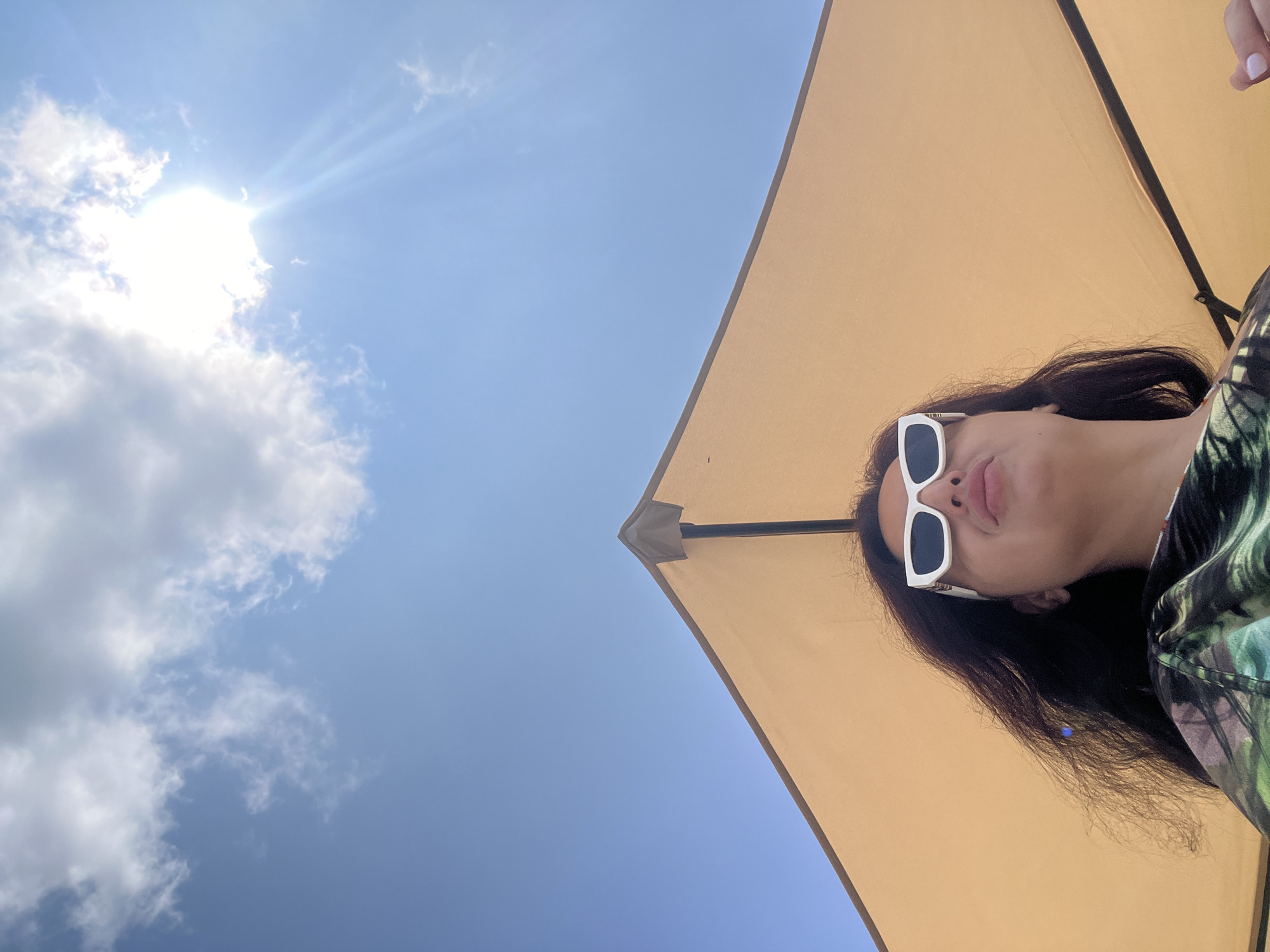In Sokhumi, Abkhazia, a woman with dark, wavy hair wears white-framed sunglasses and a green and black patterned shirt. She is positioned under a light-colored umbrella, which occupies the lower half of the frame. The sky above is a clear blue, with a few white clouds scattered across it. The sun is visible, partially obscured by clouds, casting bright rays. The image is taken from a low angle, looking up at the woman and the sky.