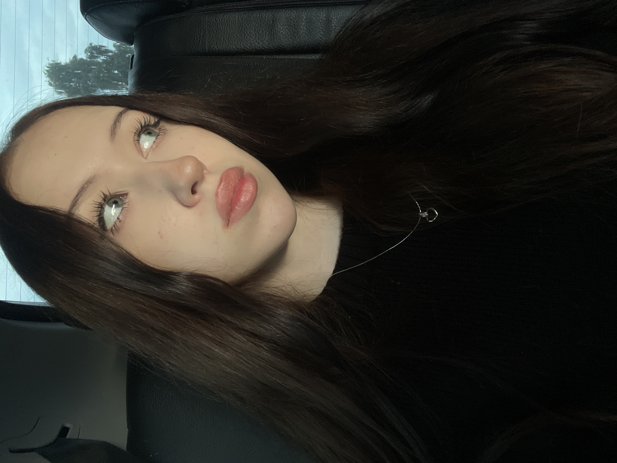 A young woman with long, dark brown hair is seated in the backseat of a vehicle. She is looking to her right, with her gaze directed away from the camera. Her lips are slightly parted, and she has a light application of lip gloss. She is wearing a black long-sleeved top, and a silver necklace with a heart-shaped pendant is visible around her neck. The background consists of a black leather car seat and a window showing a blurred outdoor scene with trees and a cloudy sky. The lighting suggests it is daytime, possibly late afternoon or evening due to the ambient light. The location is specified as Gavrilovka, Russia.