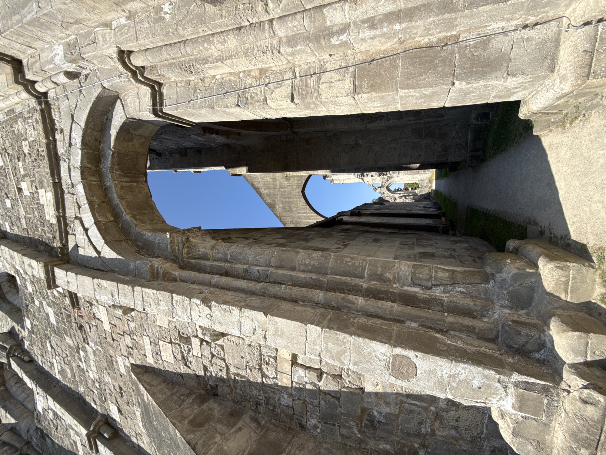 The image shows a view looking through a stone archway in the ruins of Walkenried Abbey in Walkenried, Germany. The ruins are made of large, rough-hewn stone blocks, with evidence of weathered surfaces and missing sections. The archway frames a pathway that leads further into the abbey complex, with more ruined structures visible in the distance under a clear blue sky. Sunlight casts shadows on the stone surfaces, highlighting the textures and architectural details of the ruins. There are no people or animals visible in the image.