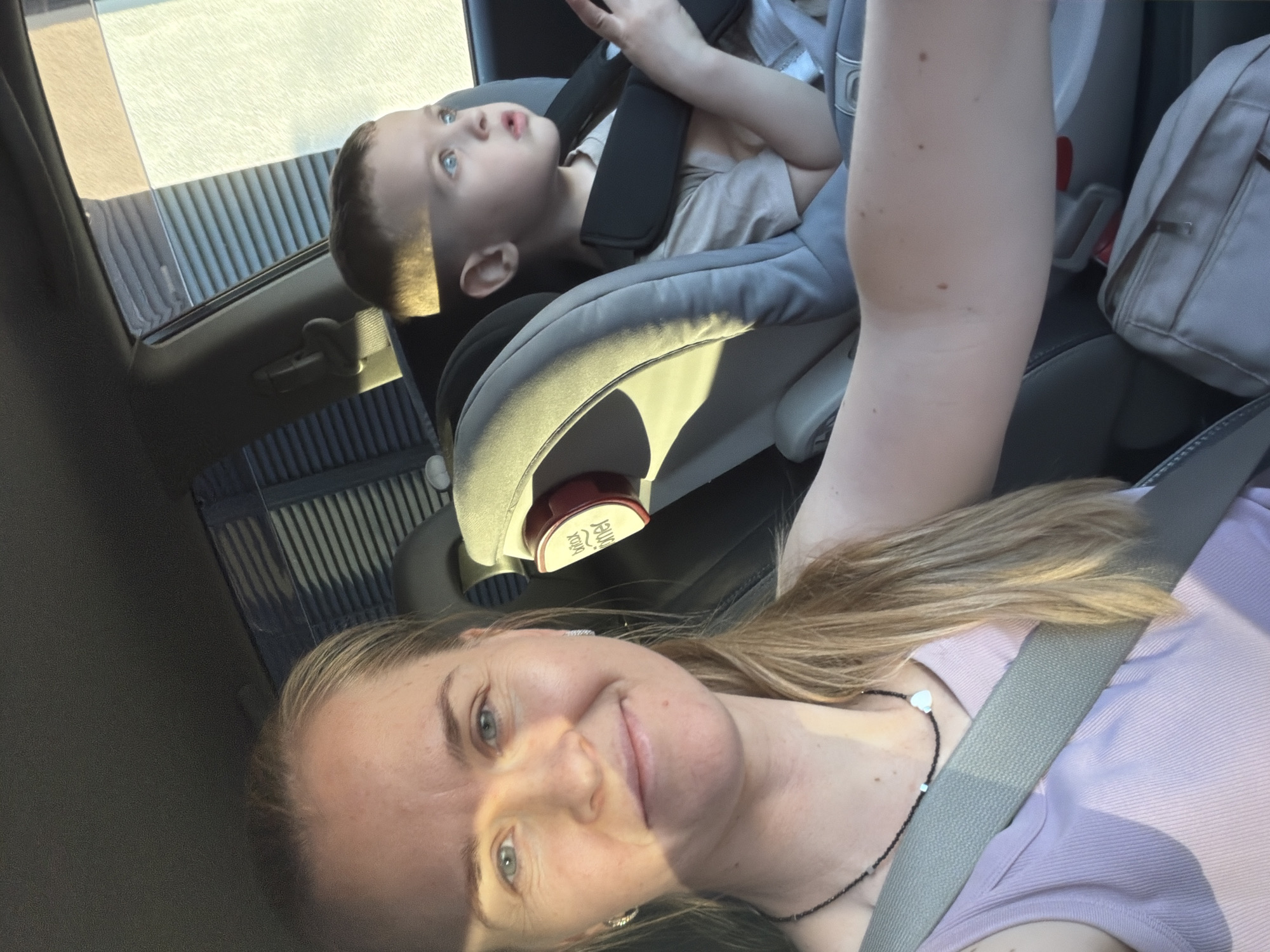 In the city of Khabarovsk, Russia, a woman and a young boy are photographed inside a car. The woman, positioned in the foreground on the left, is looking towards the camera with a slight smile. She has light-colored hair pulled back and is wearing a light purple tank top and a dark necklace. A gray seatbelt is visible across her chest. Beside her, to the right and slightly behind, a young boy is seated in a car seat. He is looking away from the camera, to his right, with a curious expression. He is wearing a light-colored t-shirt and is secured by a car seat harness. The interior of the car is visible, including the car seat and window shades. The lighting suggests it is daytime, with sunlight casting shadows.